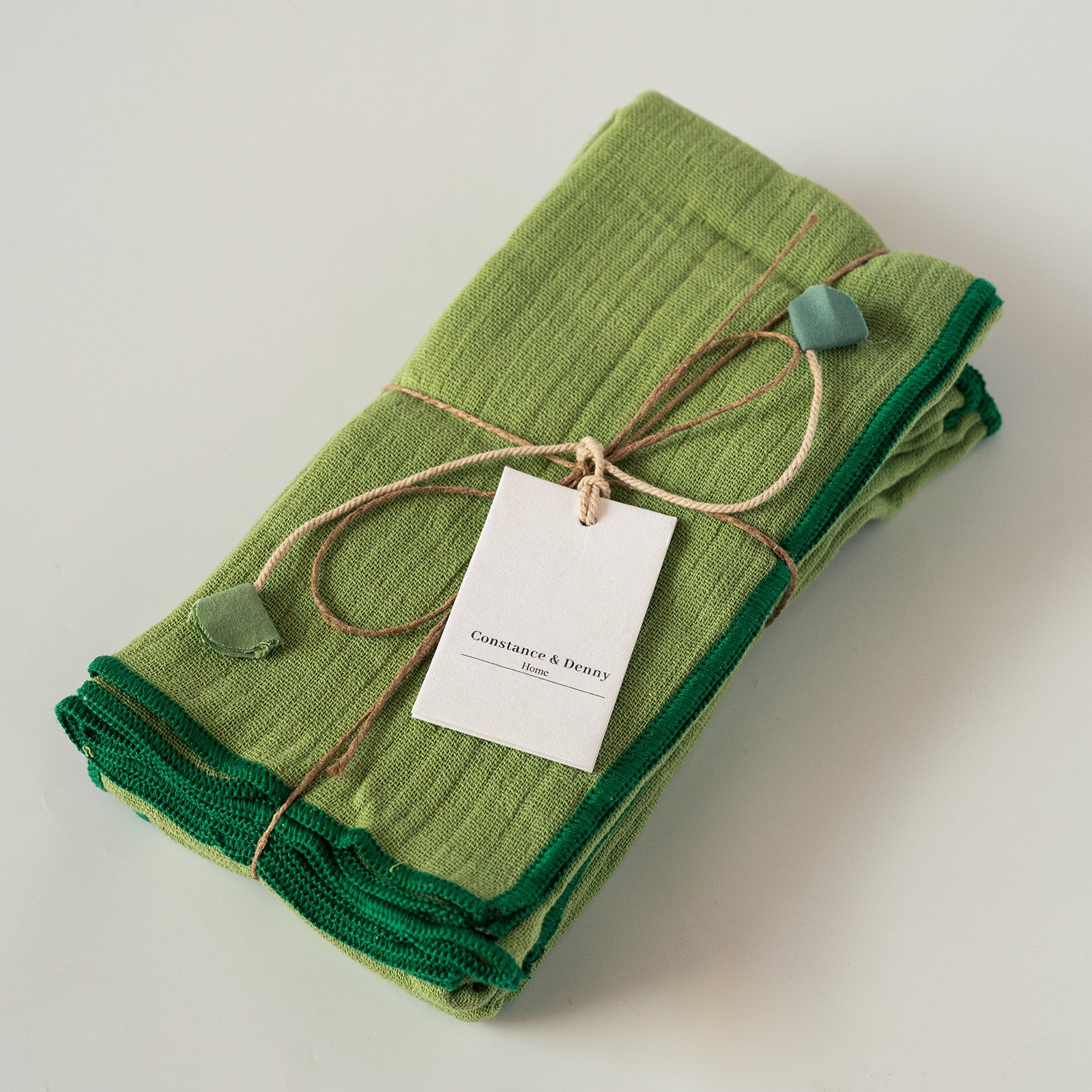A folded pair of green napkins  with dark green trim, tied with a twine string and a white paper tag that reads 'Constance & Drew'.