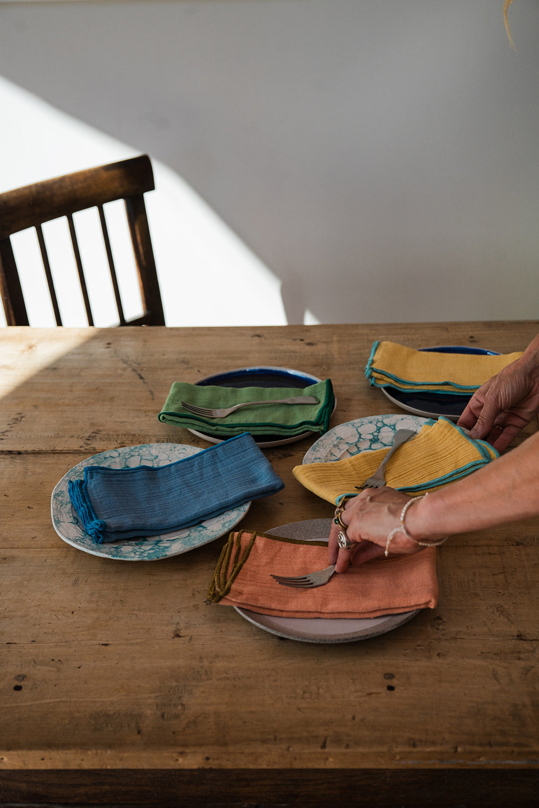 A rustic wooden dining table set with multiple place settings featuring folded linen napkins in blue, green, yellow, and orange, with a person's hands placing a fork on one of the settings.