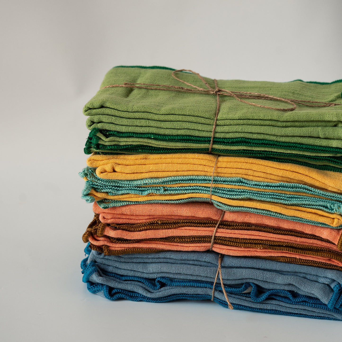 A stack of folded textured fabric napkins in various colors, including green, yellow, orange, brown, and blue, tied together with twine on a light gray background.