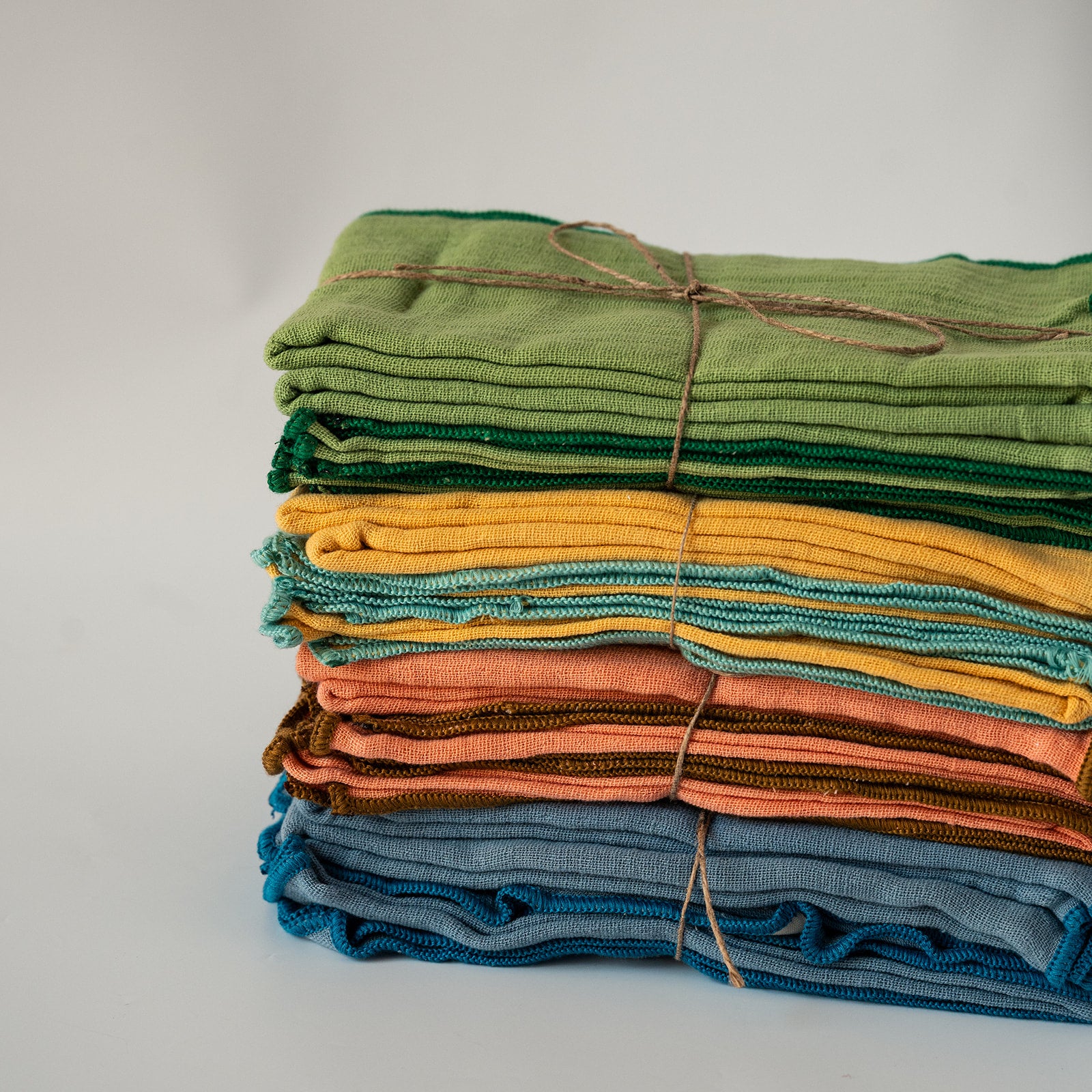 A stack of folded textured fabric napkins in various colors, including green, yellow, orange, brown, and blue, tied together with twine on a light gray background.