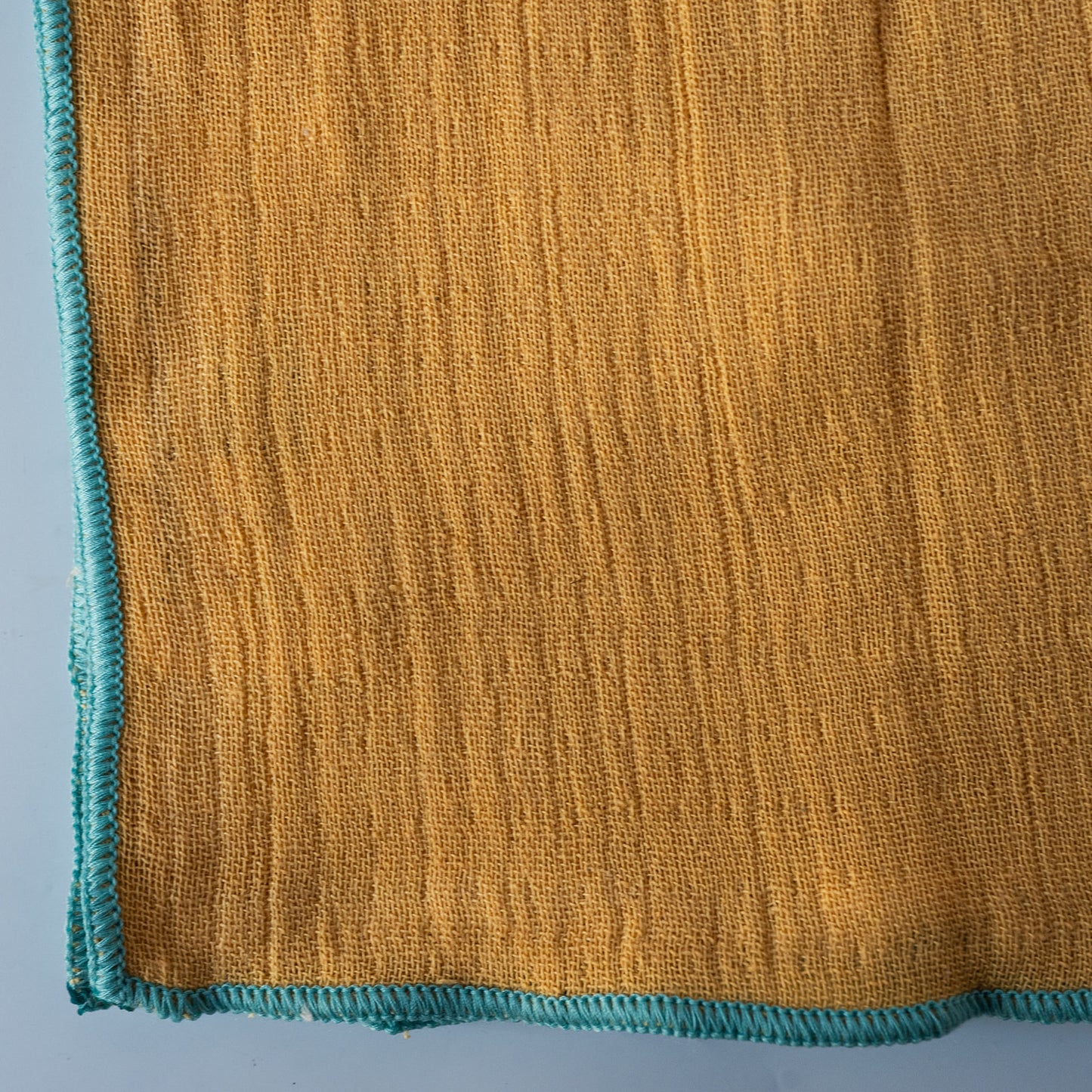 A close-up of a mustard yellow woven napkin with a textured vertical rib pattern and a contrasting teal blue stitched border.