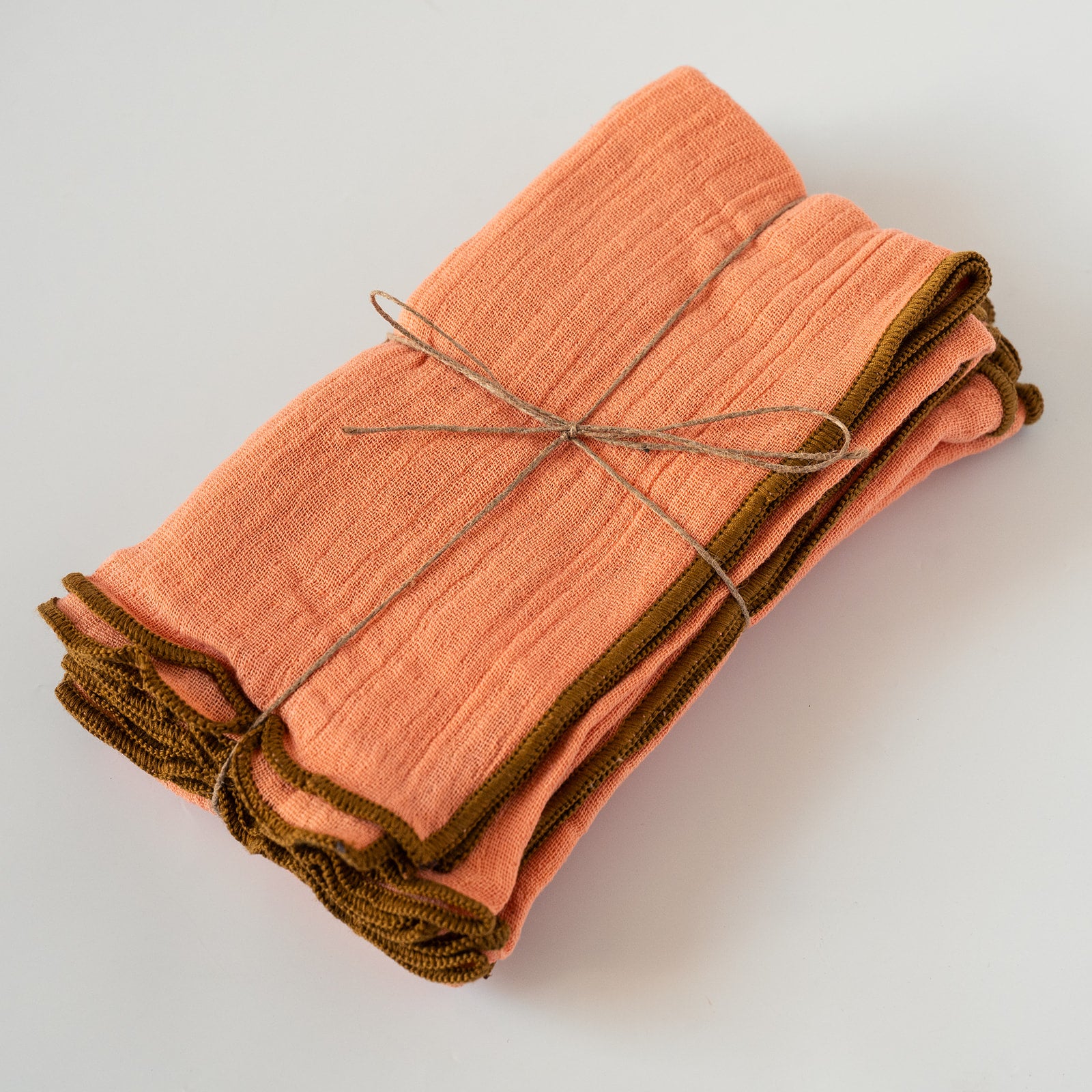 A folded set of peach-colored napkins with olive green edges, tied together with a simple twine bow on a clean white background.