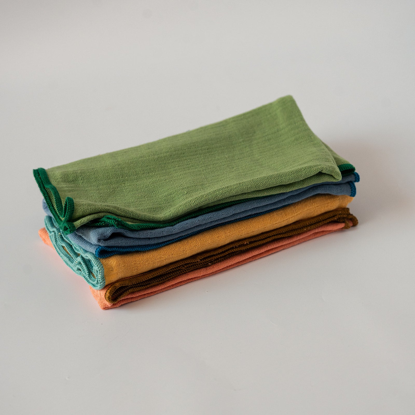 A stack of folded cotton napkins  in various earthy colors, including sage green, teal, blue, mustard yellow, brown, and terracotta, on a light gray background.