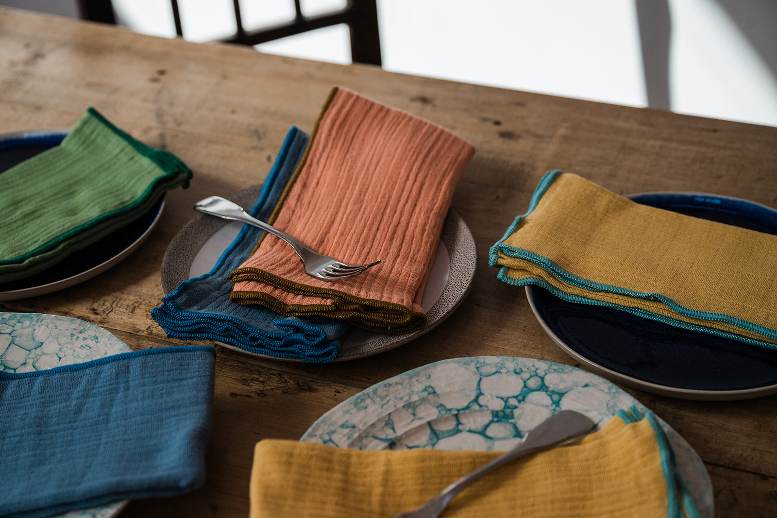 A styled dining table setting featuring textured cotton napkins in mustard yellow, teal blue, and green, folded on ceramic plates with silver cutlery on a rustic wooden table.