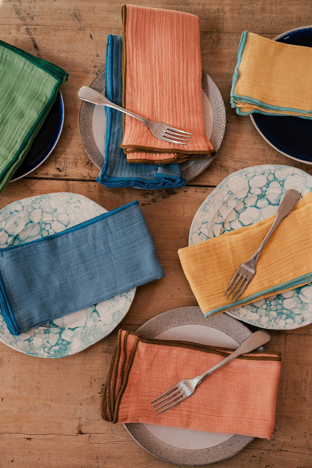 A styled flat-lay of multiple folded fabric napkins in assorted colors including blue, green, orange, and yellow, arranged on plates with forks on a rustic wooden table.