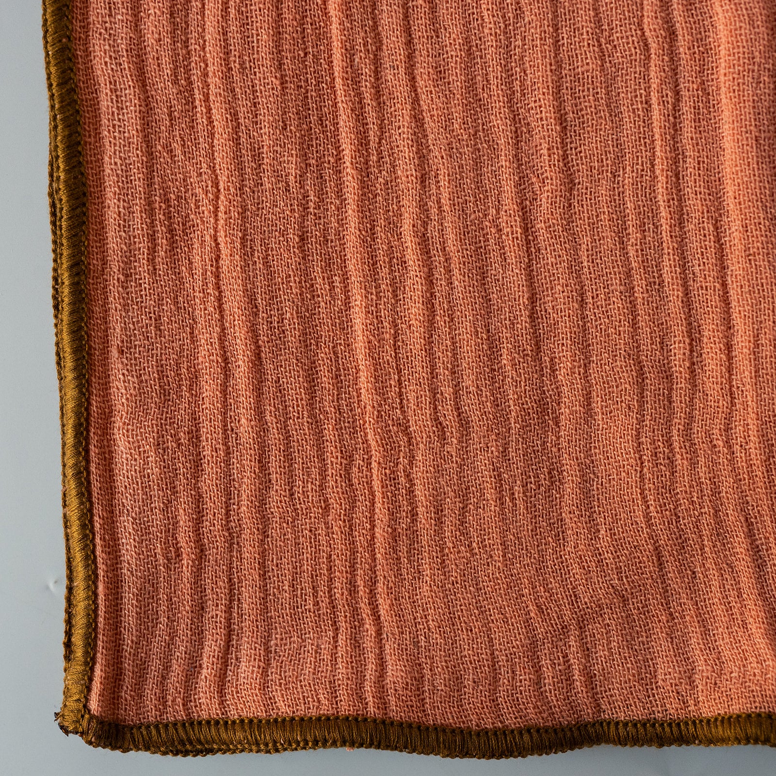 A close-up of a textured napkin in a warm terracotta orange color, showing a ribbed vertical weave and a contrasting dark brown stitched edge.