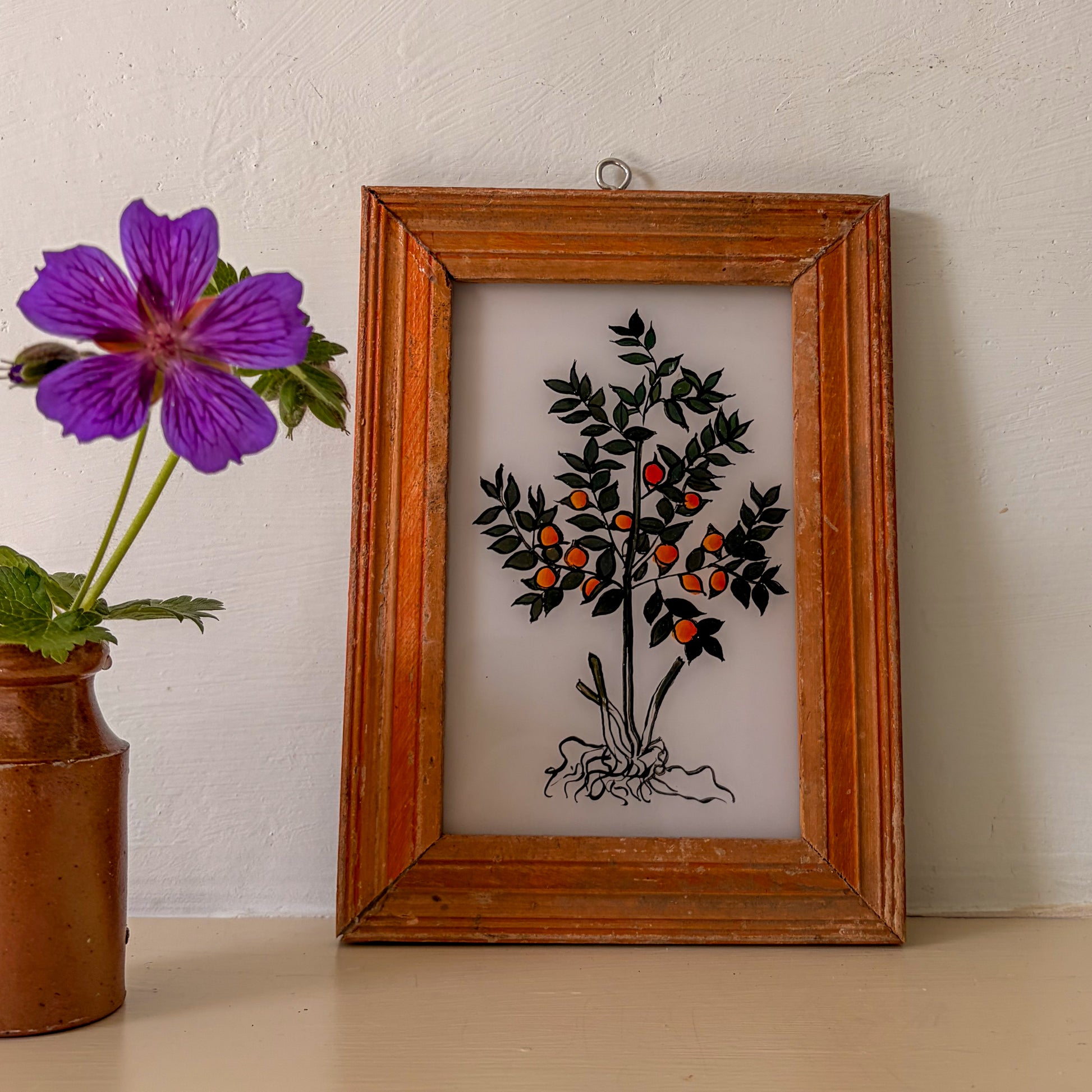 Framed artwork of a tree with fruits next to a purple flower in a brown vase on a light surface.