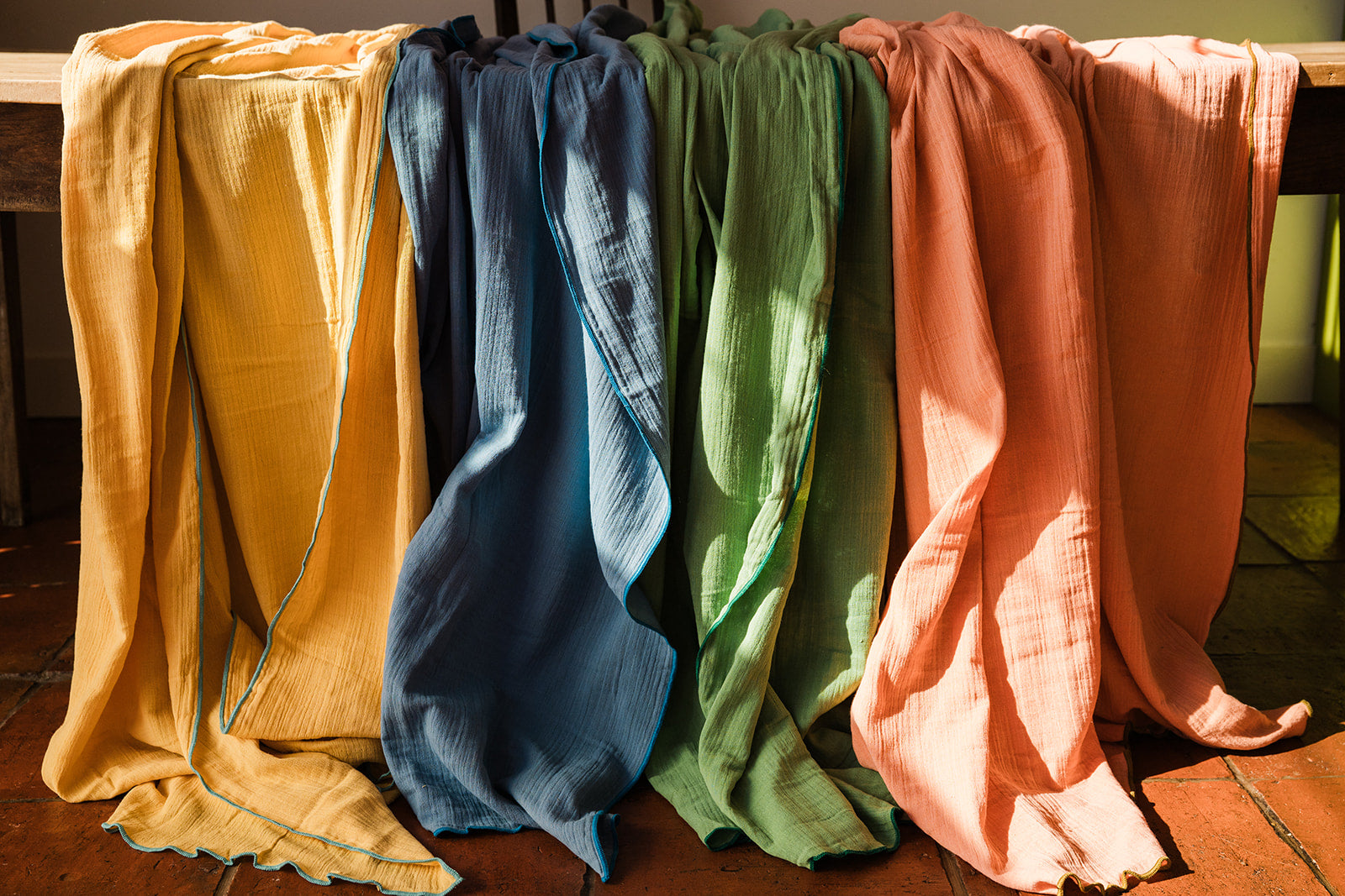 Four lightweight textured fabric cloths in mustard yellow, navy blue, sage green, and peach pink draped over a wooden table in warm natural light.