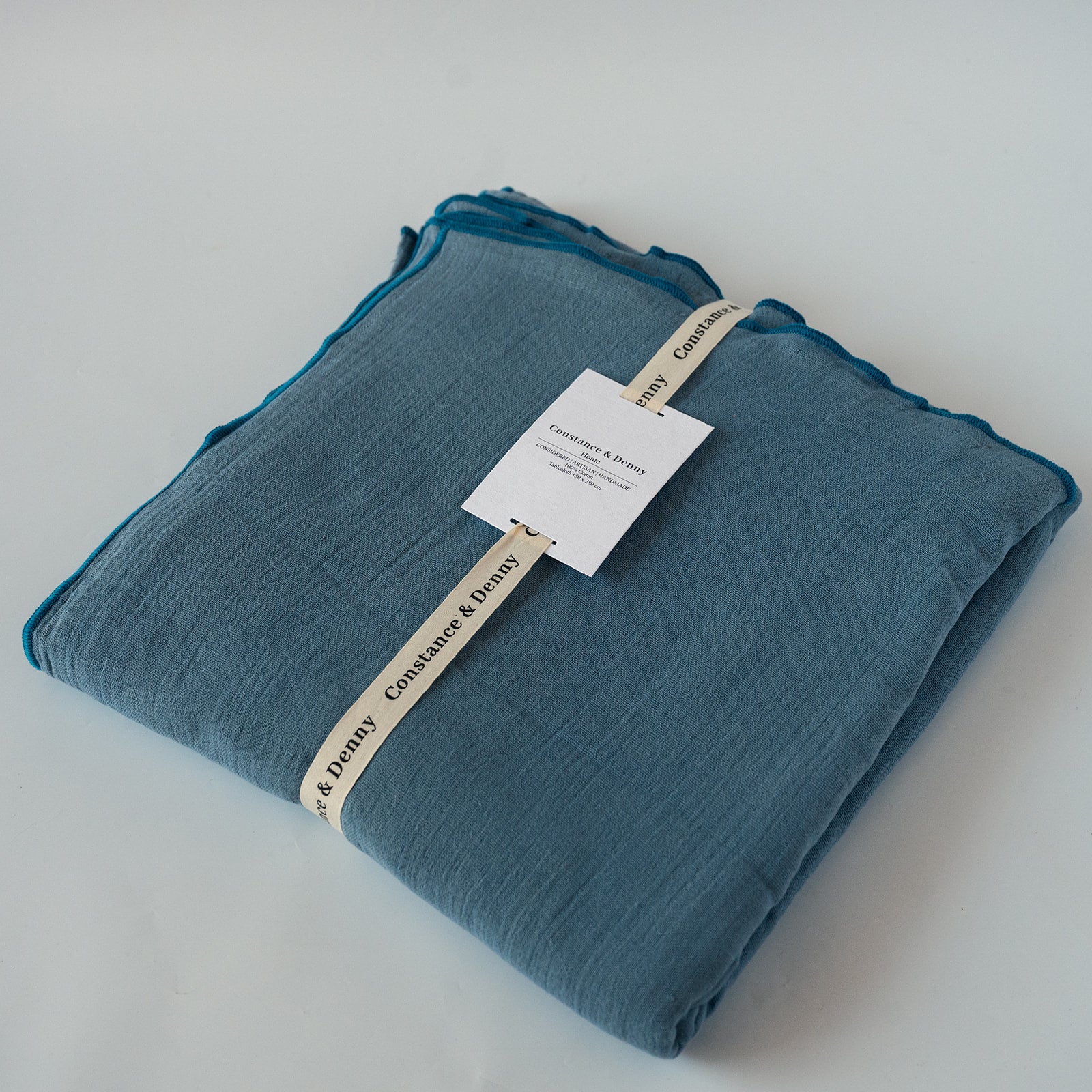 A folded dusty blue crinkle tablecloth with a white paper tag and a cream ribbon label, photographed on a light gray background.