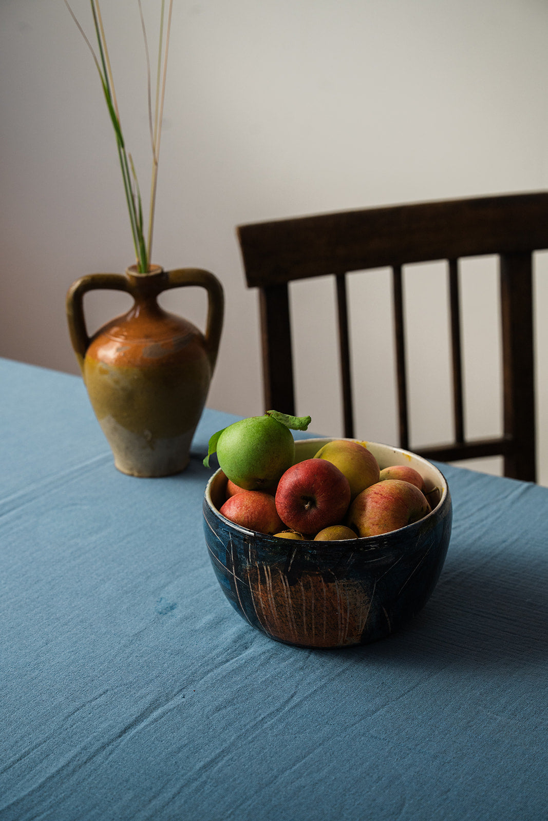 A rustic ceramic bowl filled with apples and a green pear sits on a blue tablecloth next to a wooden chair, with a matching ceramic vase holding dried stems in the background.