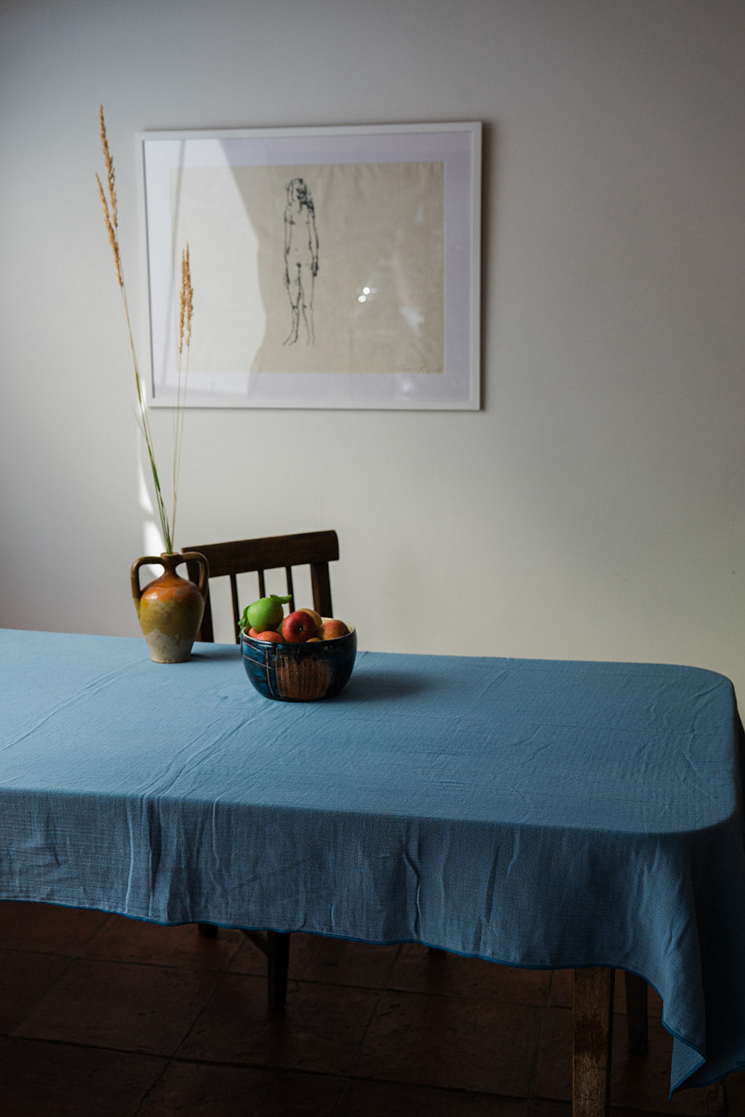 A blue tablecloth draped over a wooden dining table in a styled interior, with a ceramic vase holding dried stems and a bowl of fruit on the table. A framed art print hangs on the wall behind.