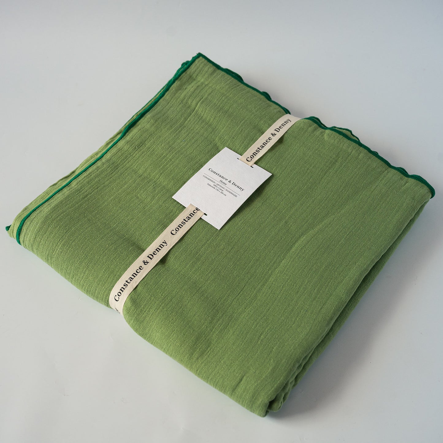 A folded sage green cotton tablecloth with a dark green border, wrapped with a cream-colored paper ribbon and a white product tag. The brand name 'Confidence & Beauty' is visible on the ribbon.