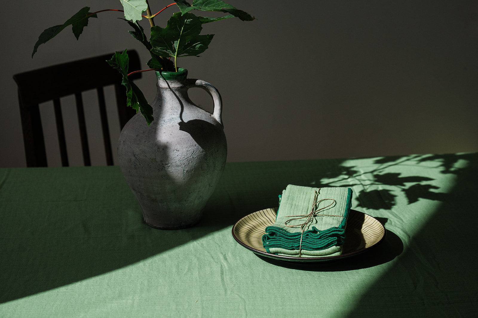A styled tabletop scene with a green tablecloth, a gray ceramic jug holding green leaves, and a stack of folded green napkins tied with twine on a plate in the foreground. Strong sunlight casts leaf shadows across the surface.