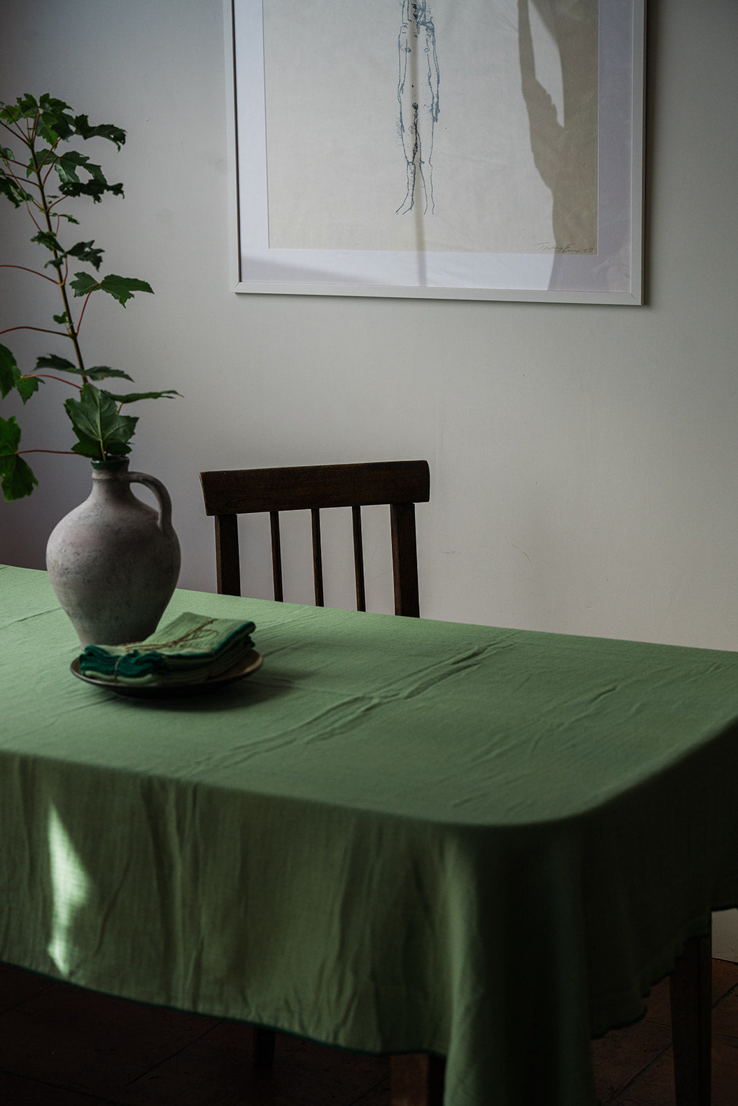 A sage green tablecloth draped over a dining table, styled with a ceramic vase containing green leaves and folded napkins. A dark wooden chair sits behind the table against a light gray wall with a framed abstract print.