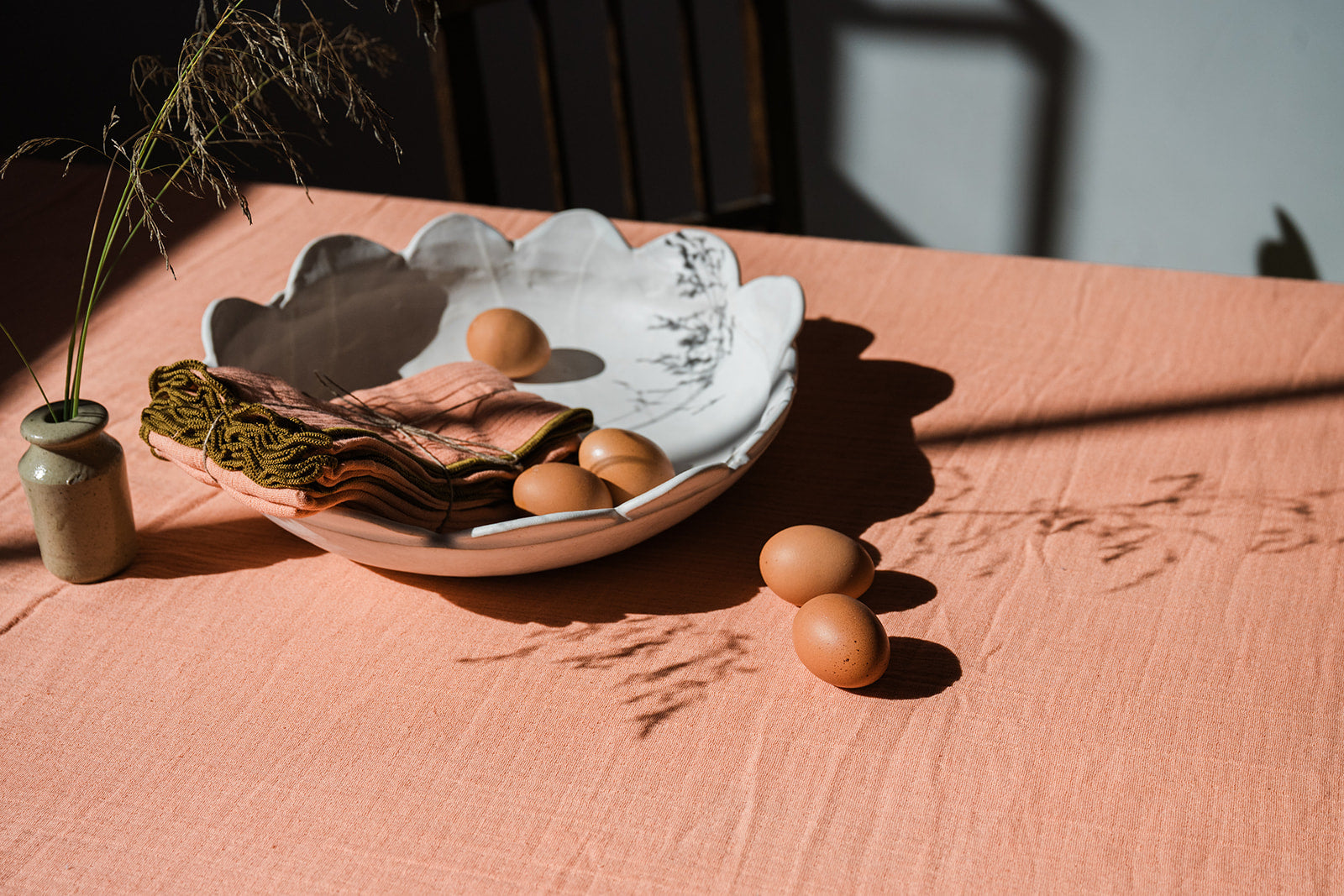 A rustic ceramic bowl with a scalloped rim, containing folded linen napkins and several brown eggs, arranged on a peach-colored tablecloth with strong sunlight and shadows.