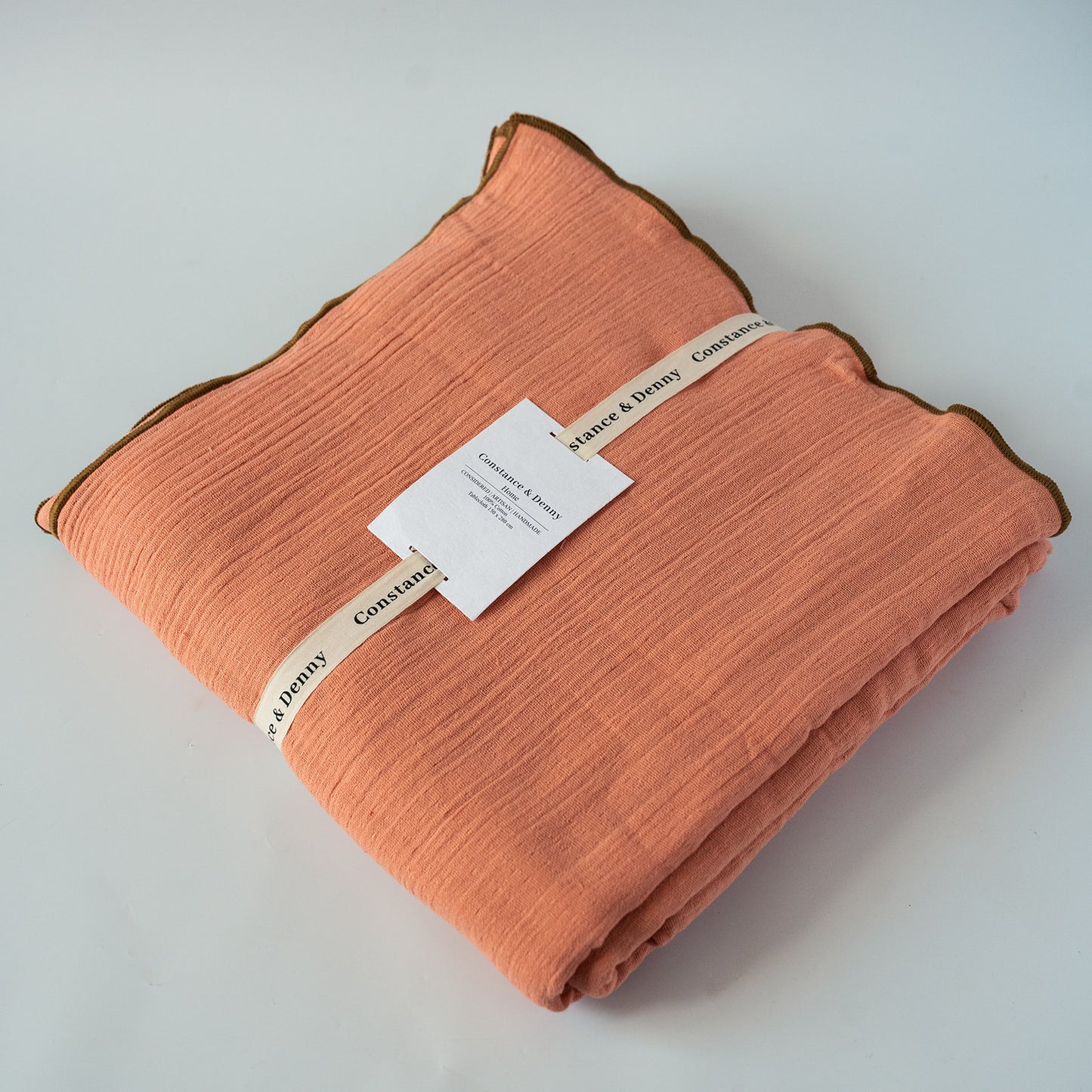 A folded peach-colored crinkle tablecloth with a brown border, wrapped with a cream paper band and a white product tag, on a light gray background.