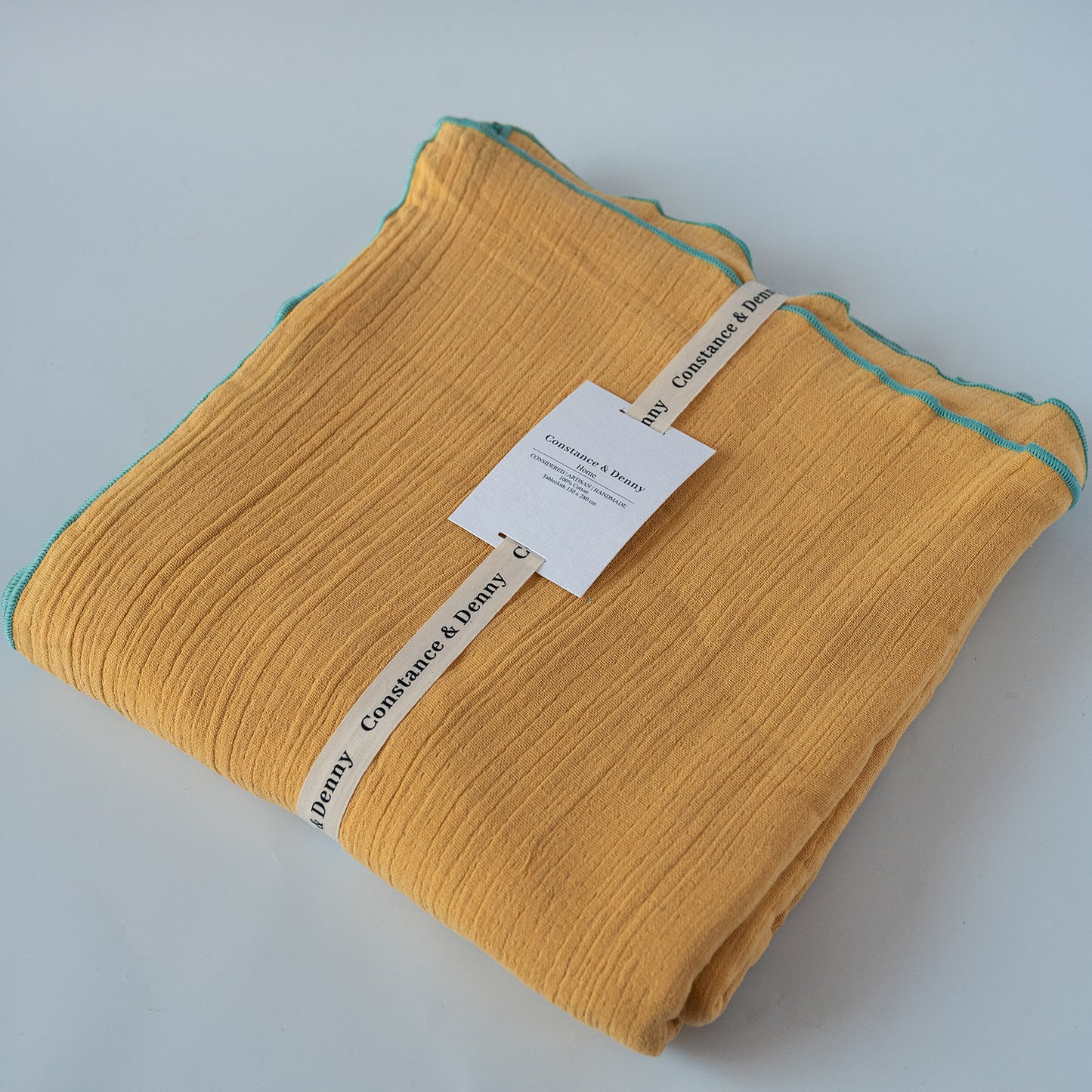 A folded mustard yellow tabelcloth with a thin teal edge trim, wrapped in a paper band labeled ' Constance & Denny' and a small product tag, on a light gray background.