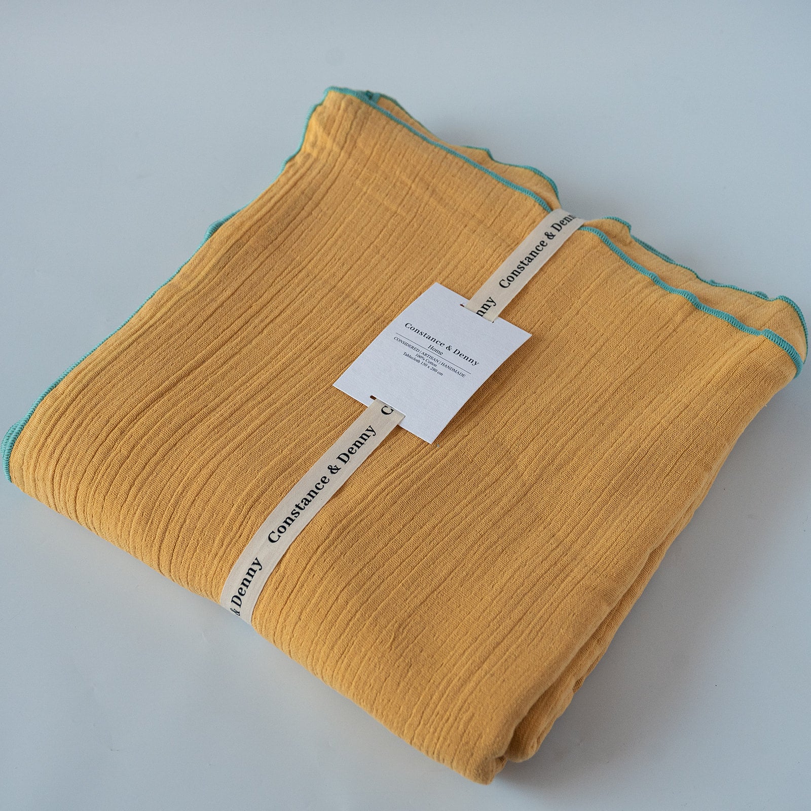 A folded mustard yellow tabelcloth with a thin teal edge trim, wrapped in a paper band labeled ' Constance & Denny' and a small product tag, on a light gray background.