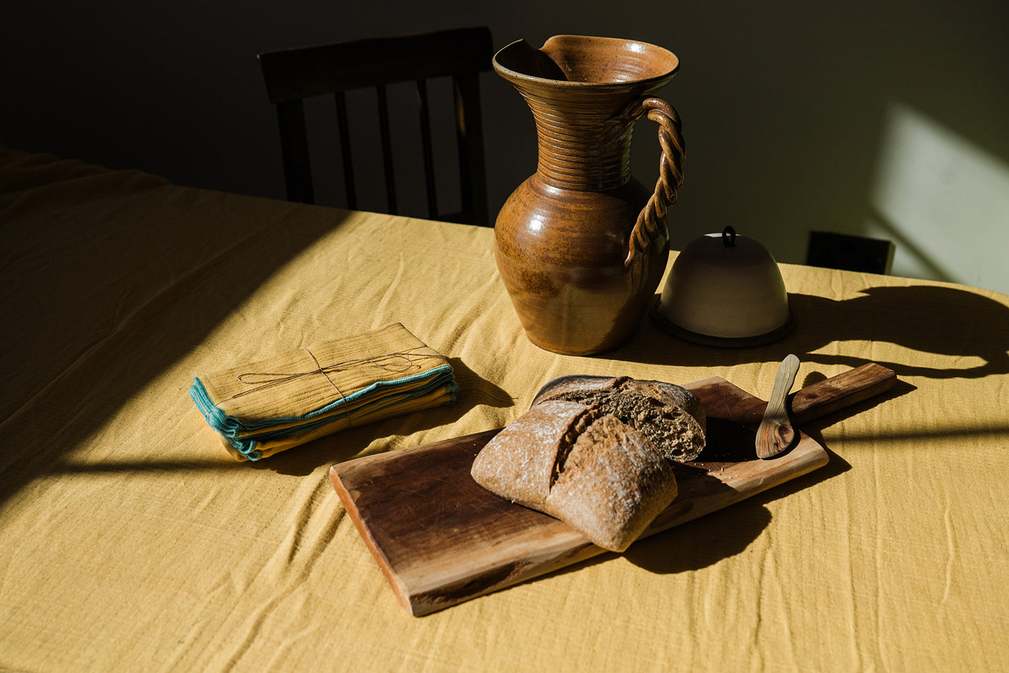A rustic still life scene on a yellow tablecloth with a wooden cutting board holding a loaf of bread and a small wooden utensil, a folded cloth with string on the left, and a brown ceramic pitcher and small white container behind it. Strong sunlight casts deep shadows across the scene.