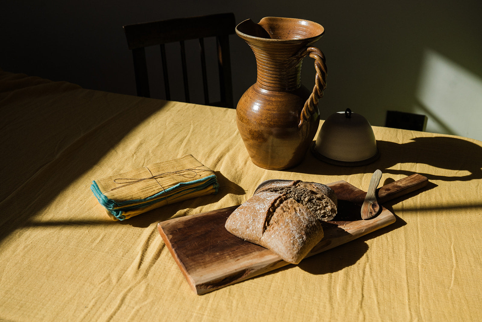 A rustic still life scene on a yellow tablecloth with a wooden cutting board holding a loaf of bread and a small wooden utensil, a folded cloth with string on the left, and a brown ceramic pitcher and small white container behind it. Strong sunlight casts deep shadows across the scene.