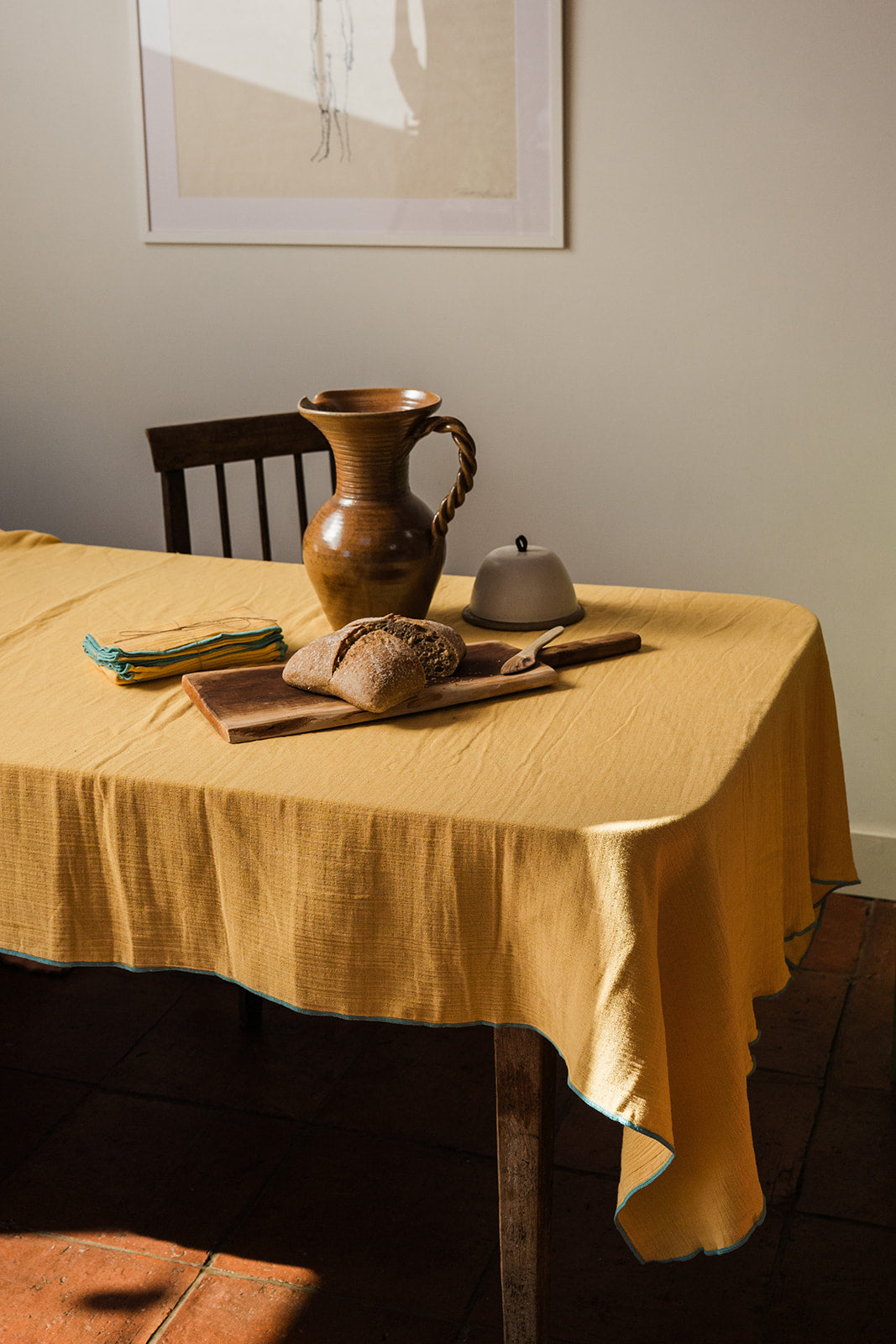 A mustard yellow linen tablecloth draped over a wooden dining table in a warm, rustic room setting, styled with a ceramic pitcher, bread, and a small bowl.