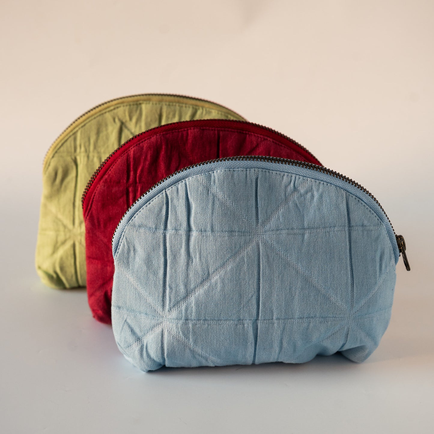 Three quilted zippered cosmetic pouches in light blue, red, and olive green, arranged in a row on a light beige background.