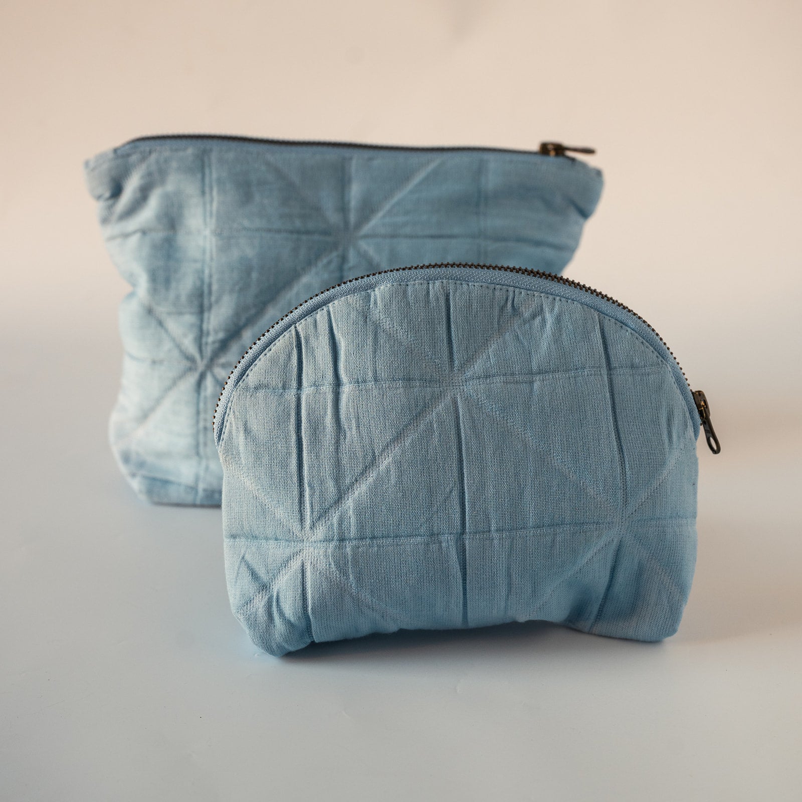 Two quilted fabric zippered pouches in a light blue denim-like material, with one larger rectangular pouch standing behind a smaller oval pouch in the foreground, photographed on a plain light background.