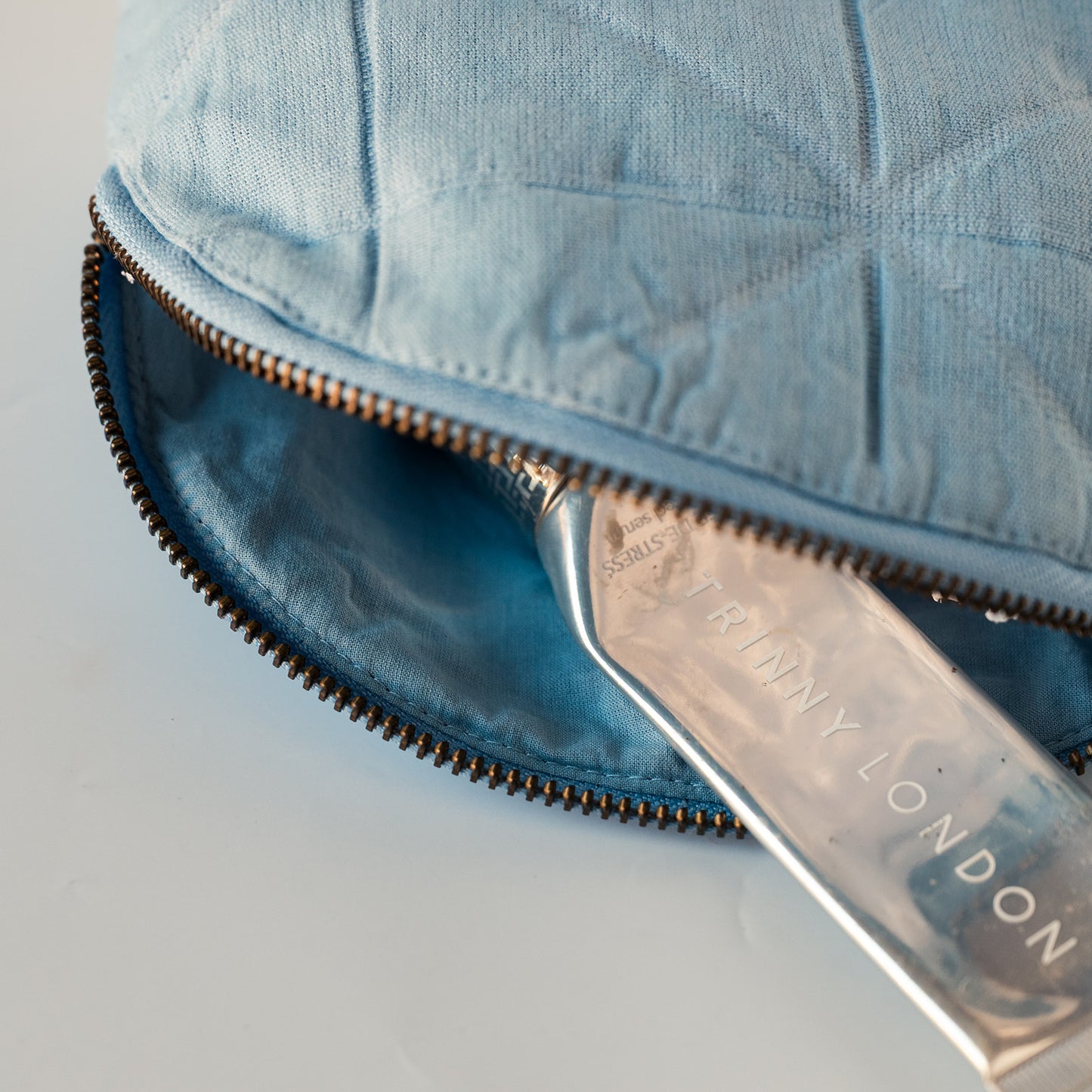 A close-up of a light blue quilted fabric pouch or cosmetic bag with a dark zipper, showing a silver metal tool with 'TRINNY LONDON' printed on it resting inside.