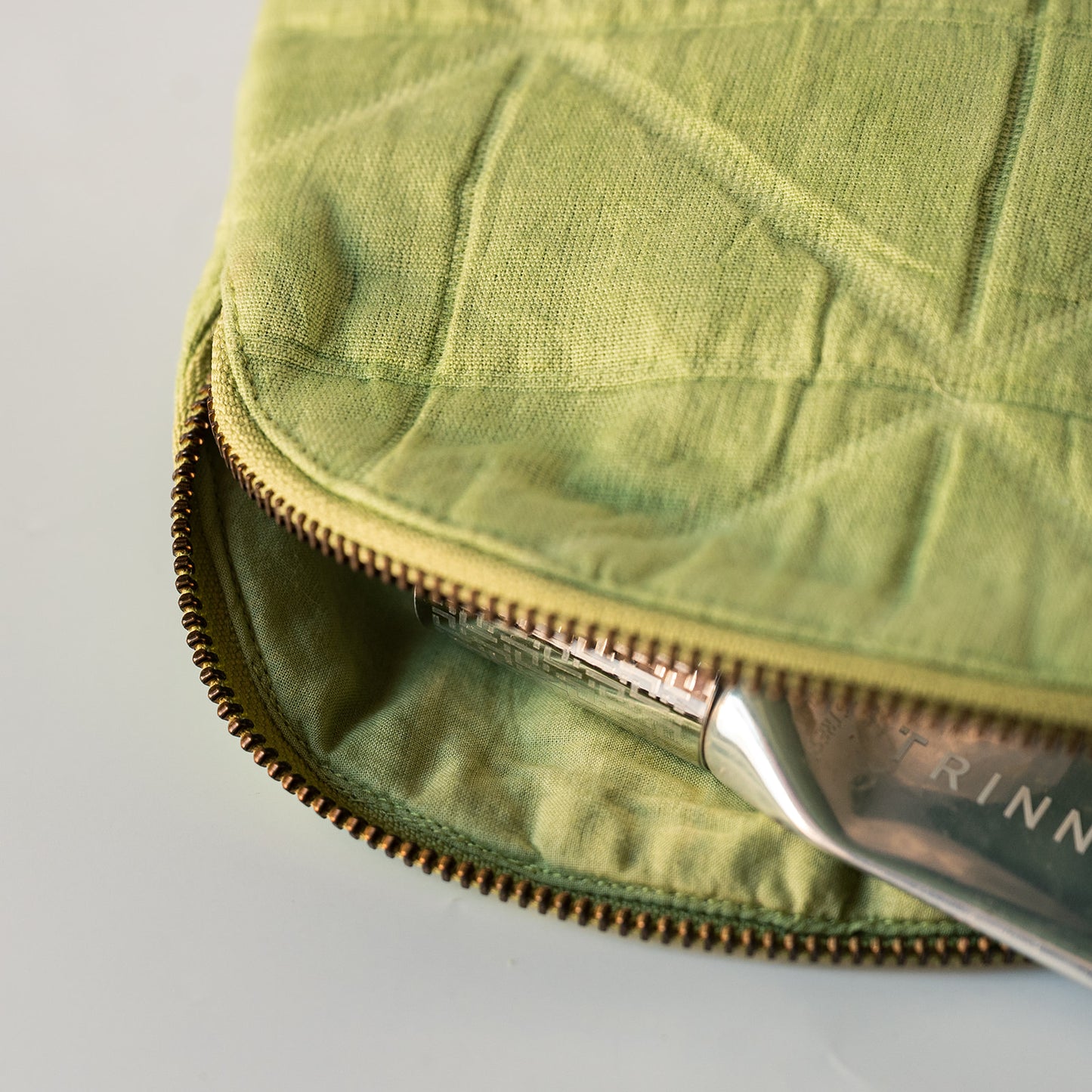 Close-up of a quilted olive green fabric pouch with a brass zipper, showing a silver metal tool partially inserted inside.
