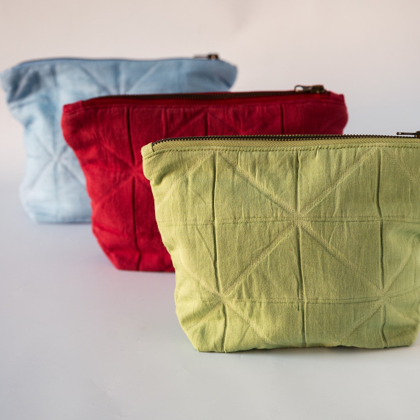Three quilted fabric zippered pouches in light blue, red, and light green, arranged diagonally on a white surface against a white background.