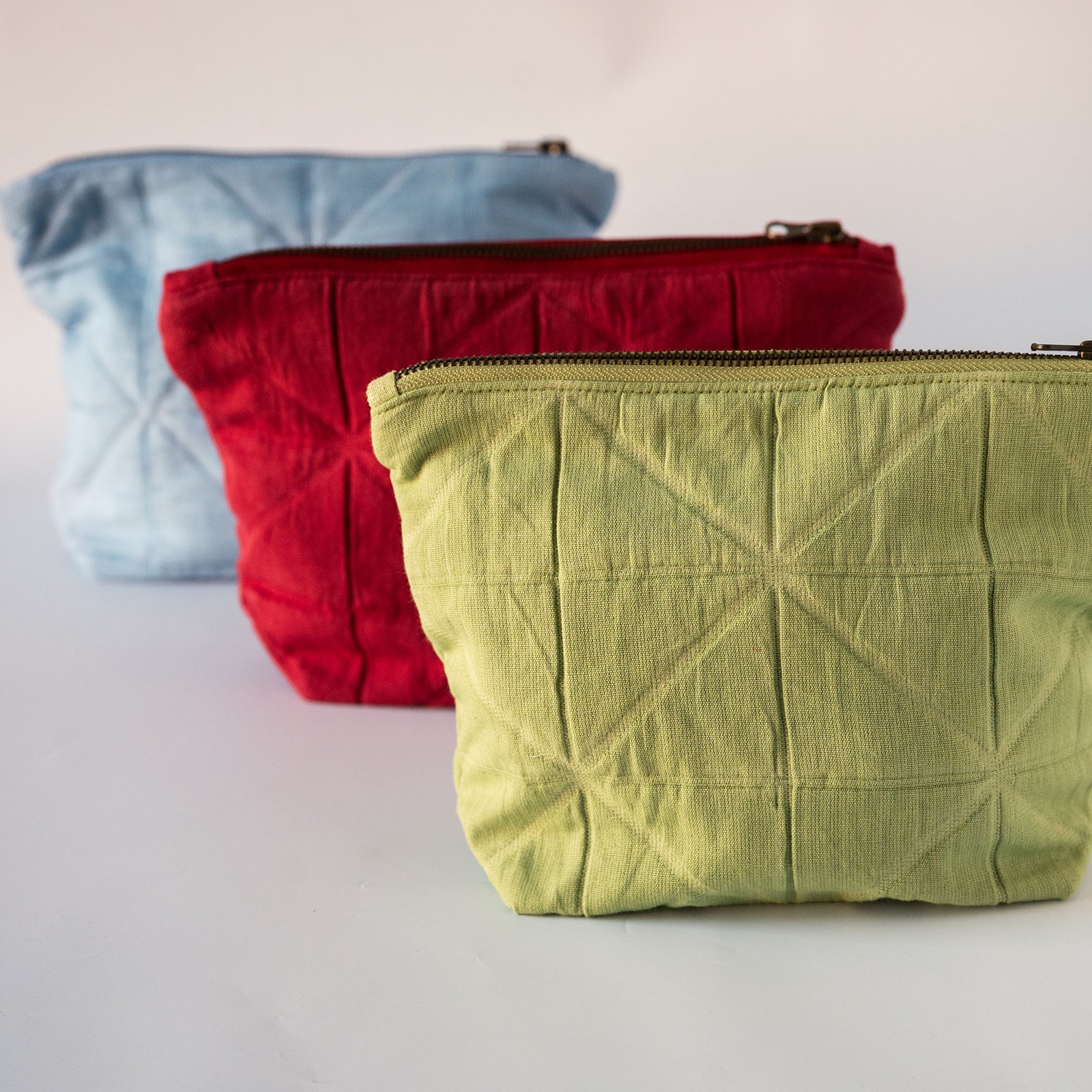 Three quilted fabric zippered pouches in light blue, red, and light green, arranged diagonally on a white surface against a white background.