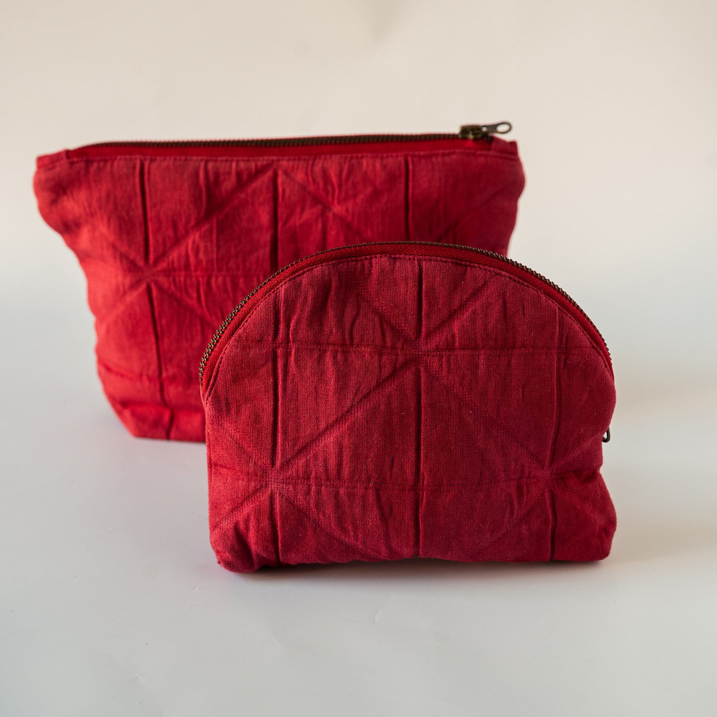 Two quilted fabric zippered pouches in a deep red color, one rectangular and one semi-circular, displayed on a light off-white background.