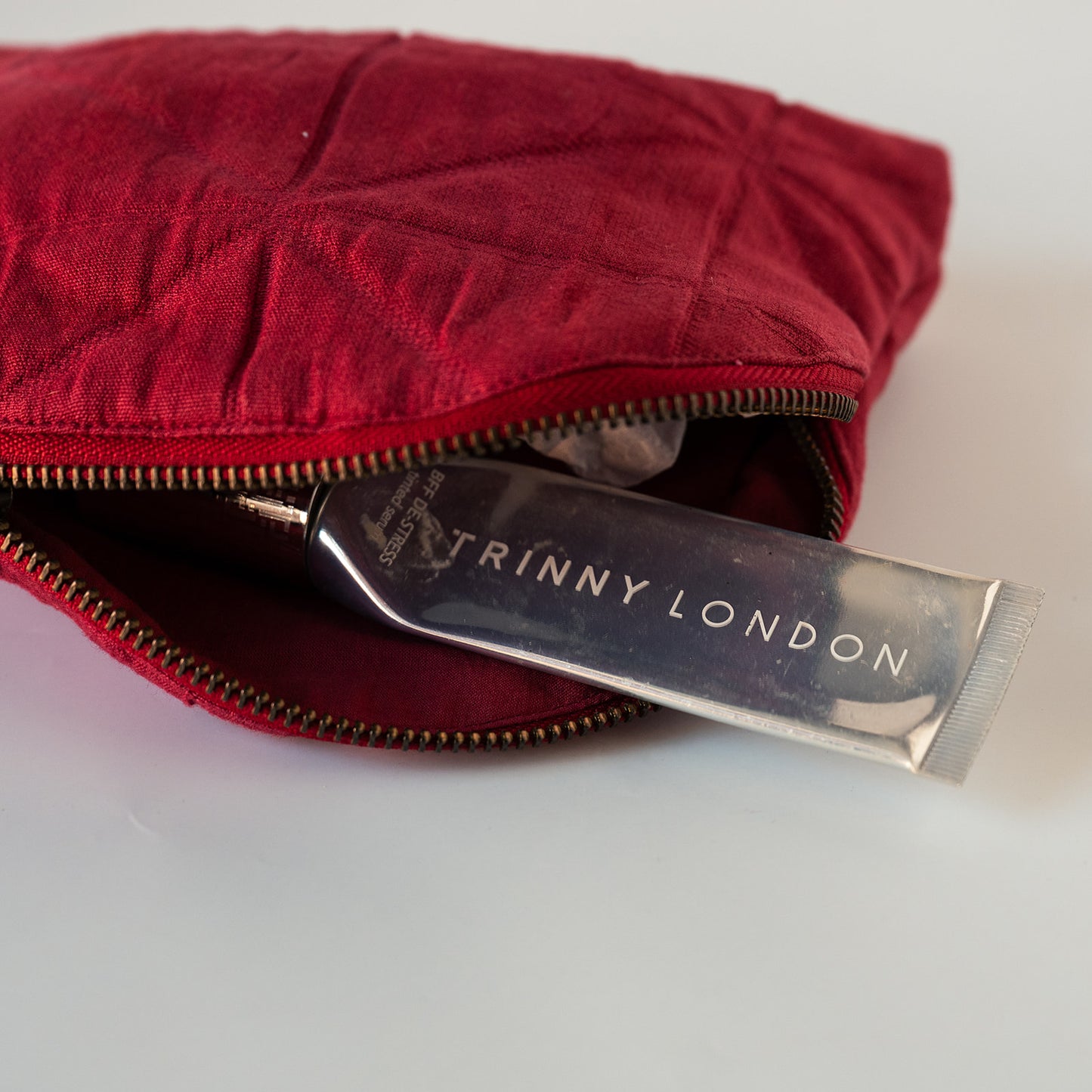 A close-up of a red quilted cosmetic pouch with a gold zipper, partially open to reveal a silver tube of Triny London cream inside, set against a plain white background.