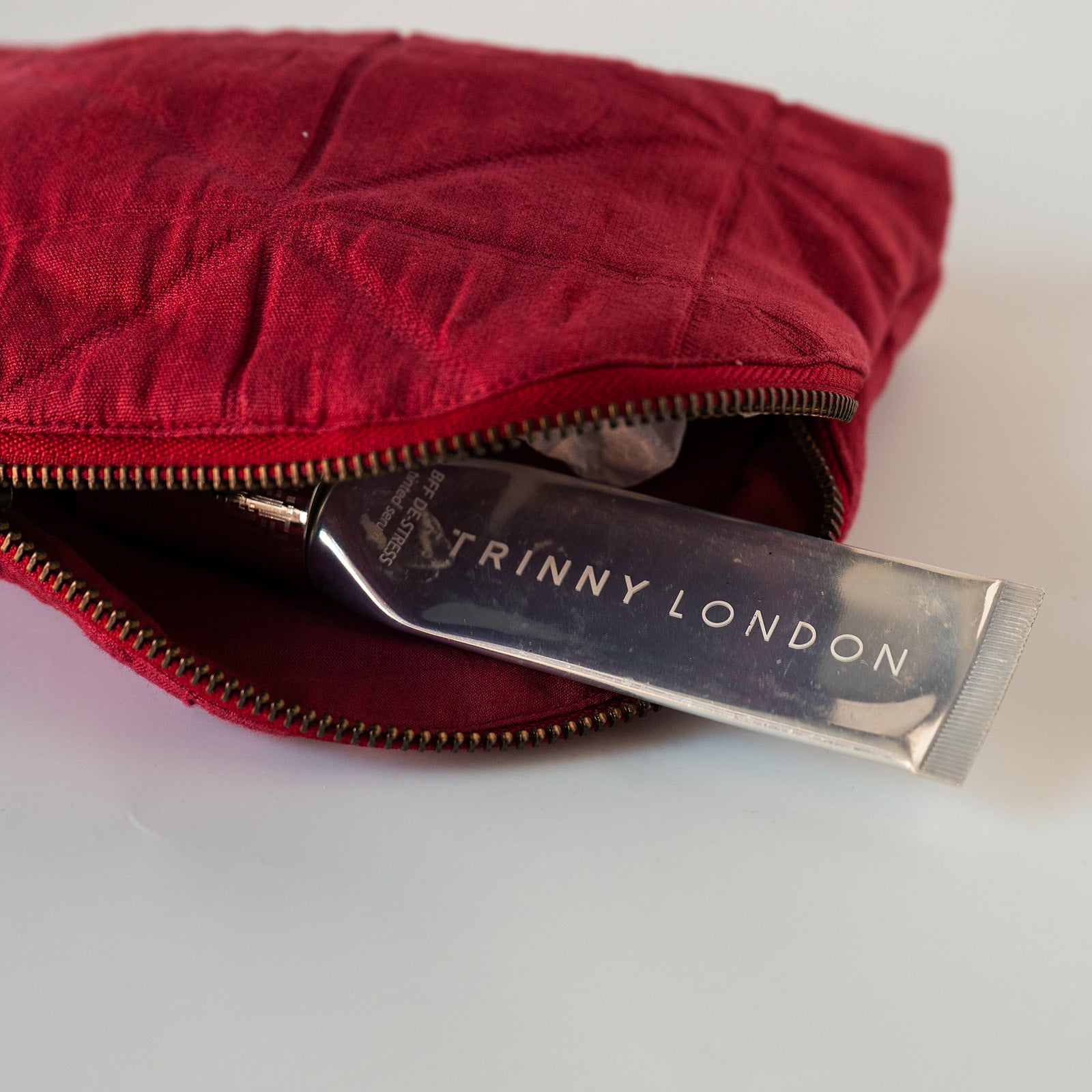 A close-up of a red quilted cosmetic pouch with a gold zipper, partially open to reveal a silver tube of Triny London cream inside, set against a plain white background.