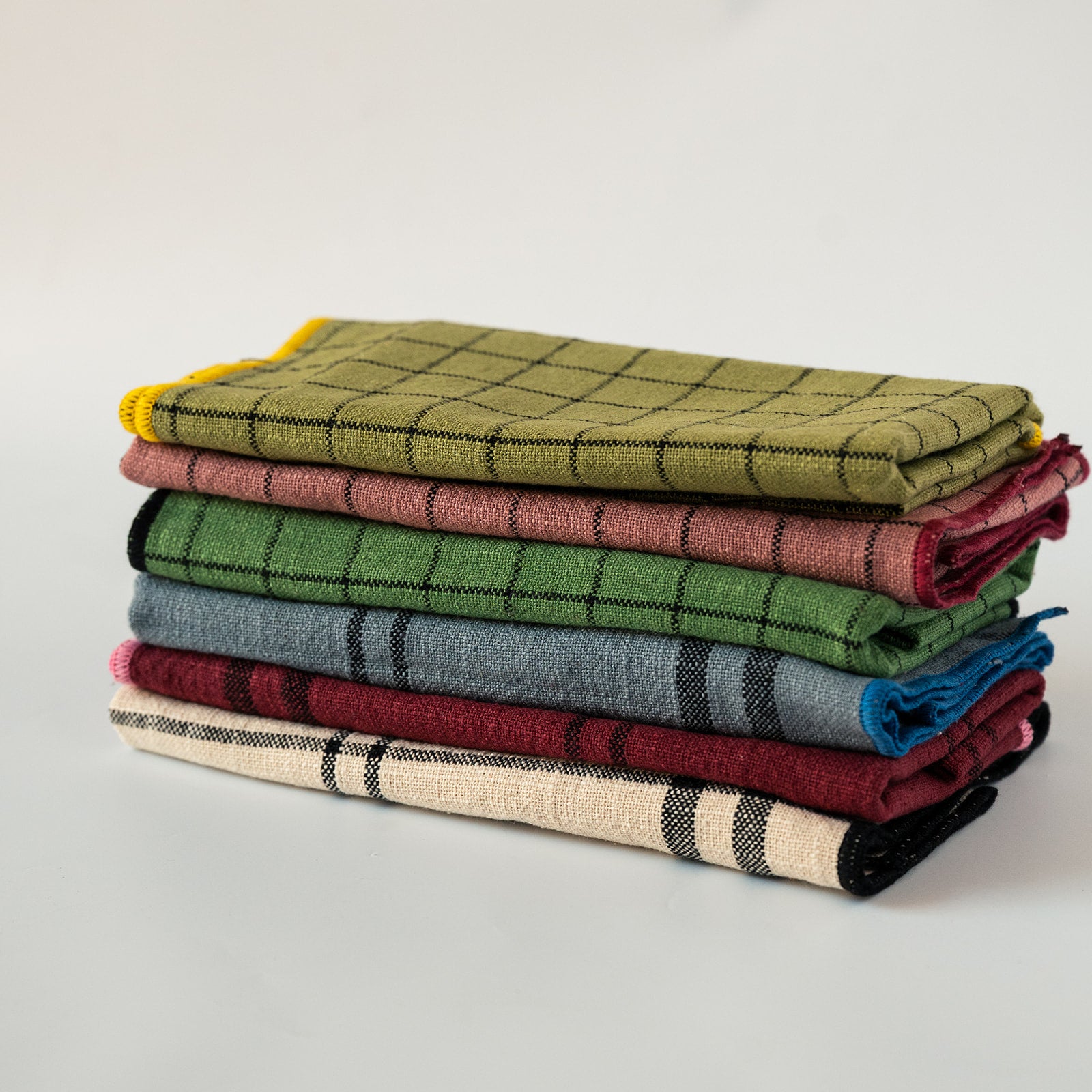 A stack of six folded plaid blankets in various colors, including olive green, dusty rose, forest green, slate blue, burgundy, and cream, set against a plain light gray background.