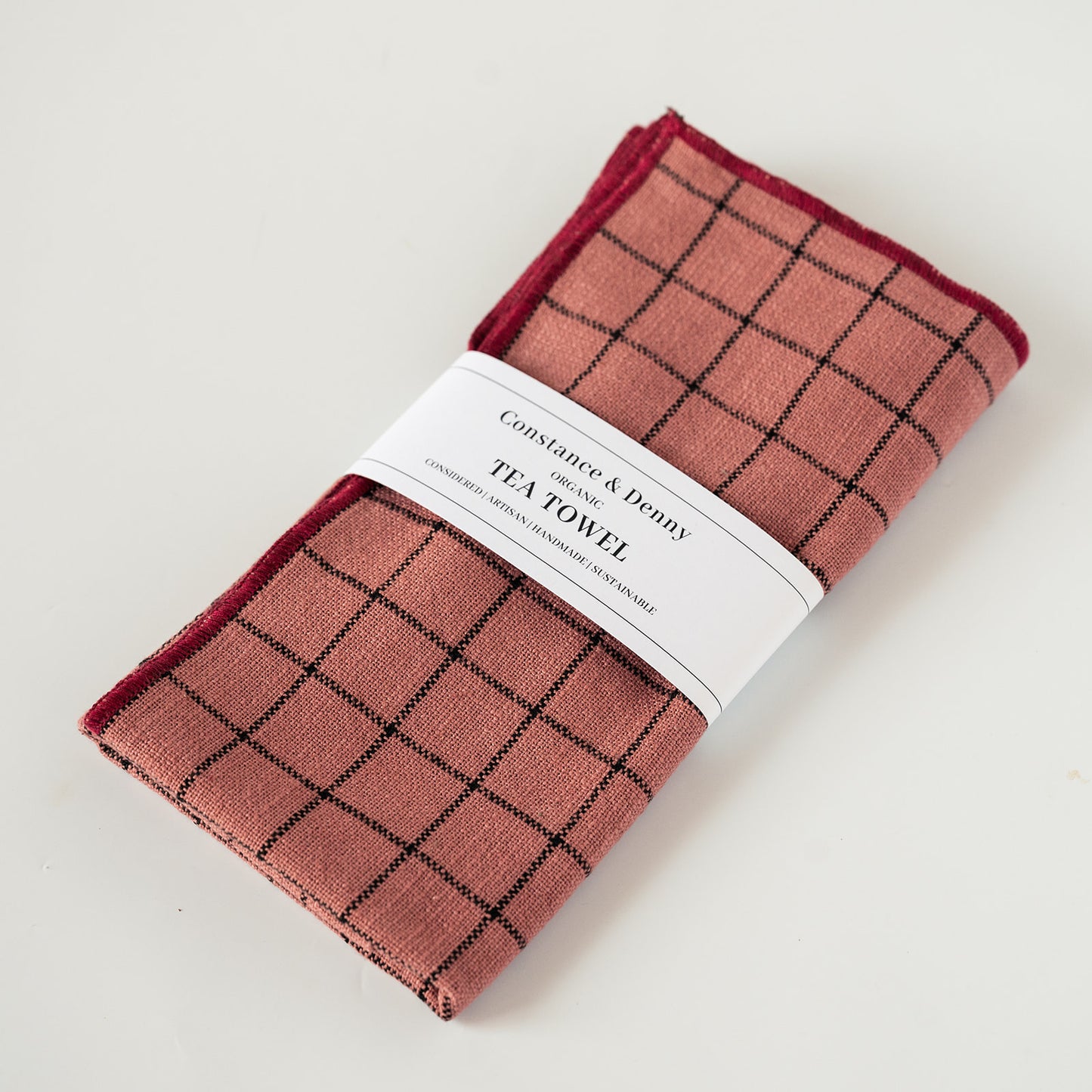 A folded tea towel with a dusty rose and black plaid pattern, wrapped in a white paper band that reads 'Constance & Denny TEA TOWEL'.