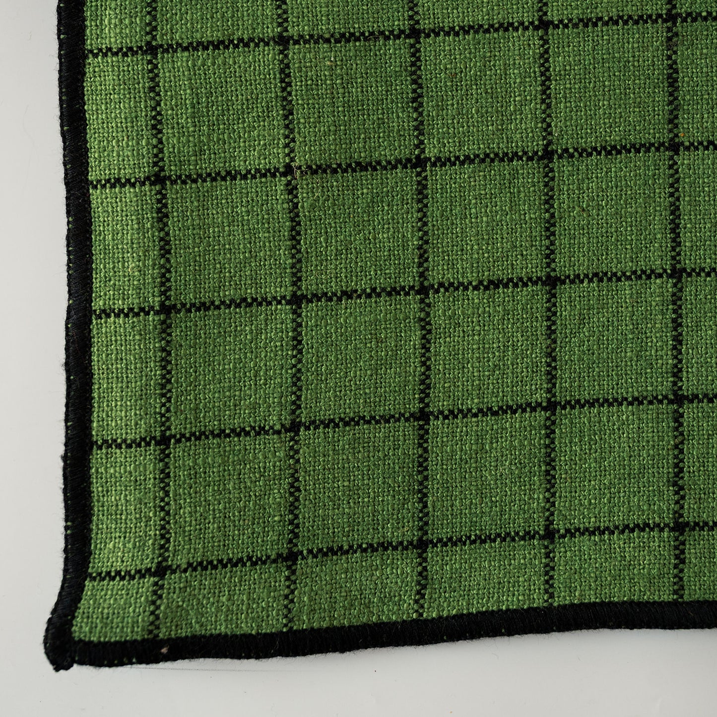 A close-up of a green woven textile with a black grid pattern and black stitched border, shown on a white background.