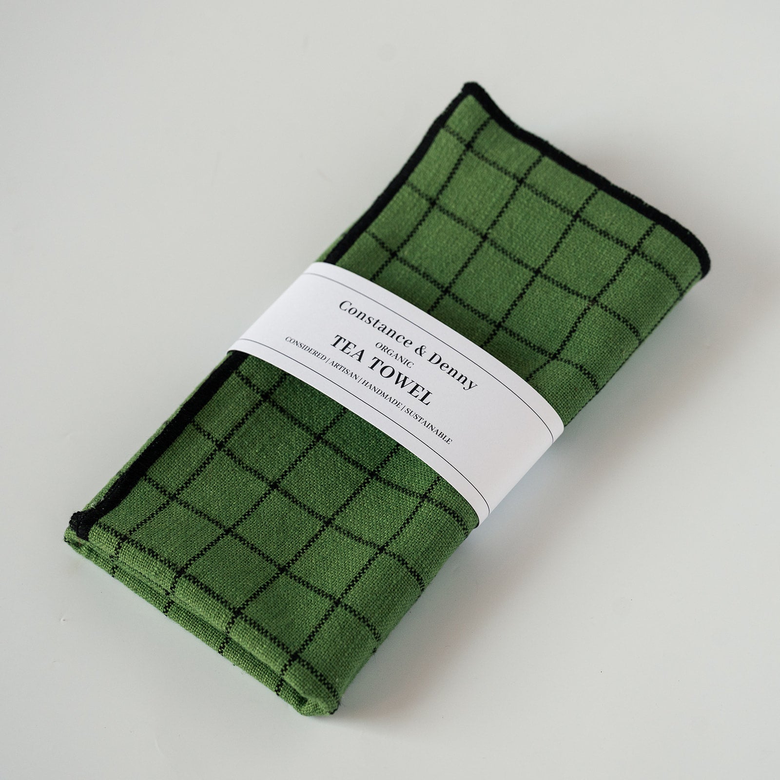 A folded green tea towel with a black windowpane plaid pattern, wrapped in a white paper band that reads 'Constance & Deny' and 'TEA TOWEL'.