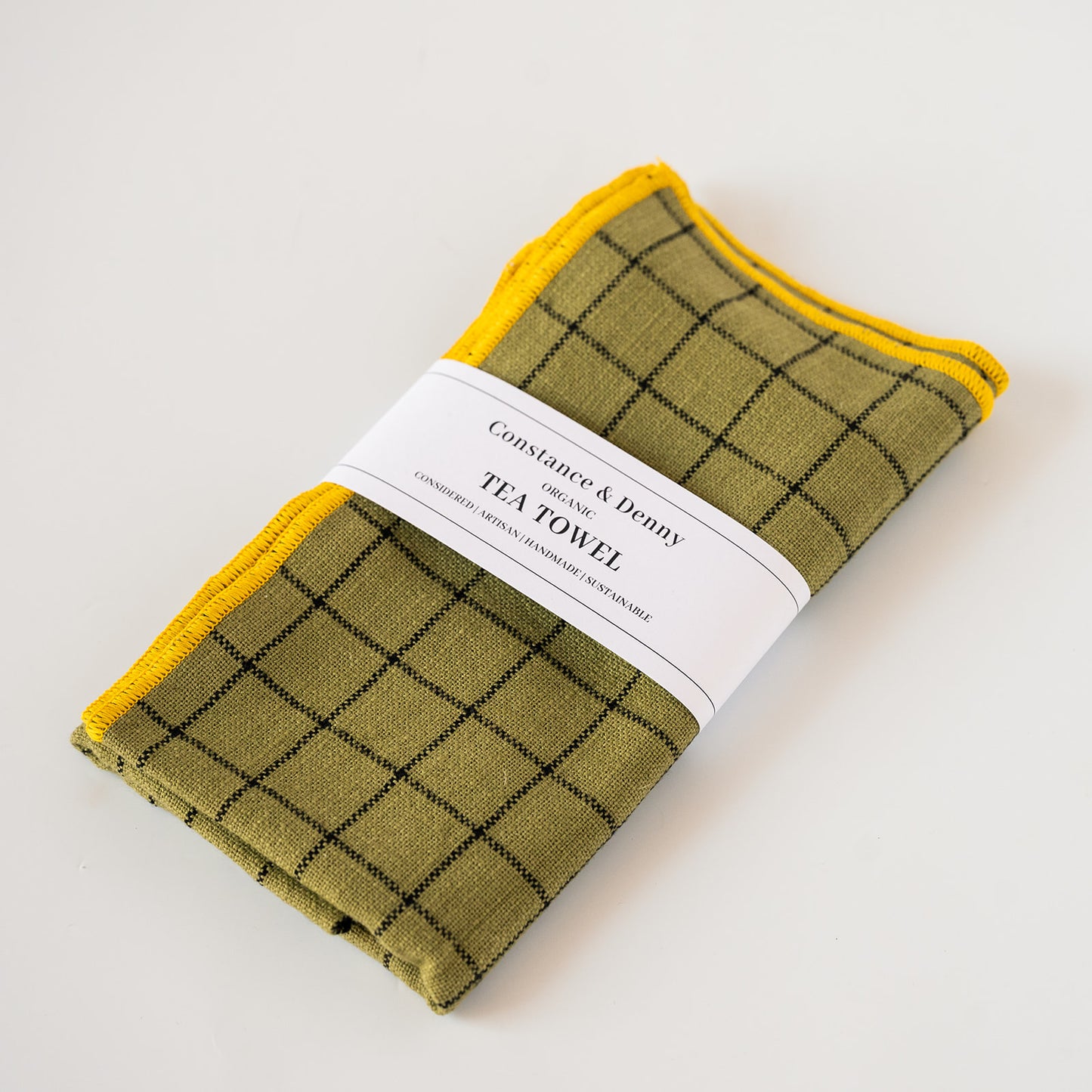 A folded tea towel with an olive green and black windowpane plaid pattern and a bright yellow border, wrapped in a white paper band labeled 'Constance & Benny Tea Towel'.