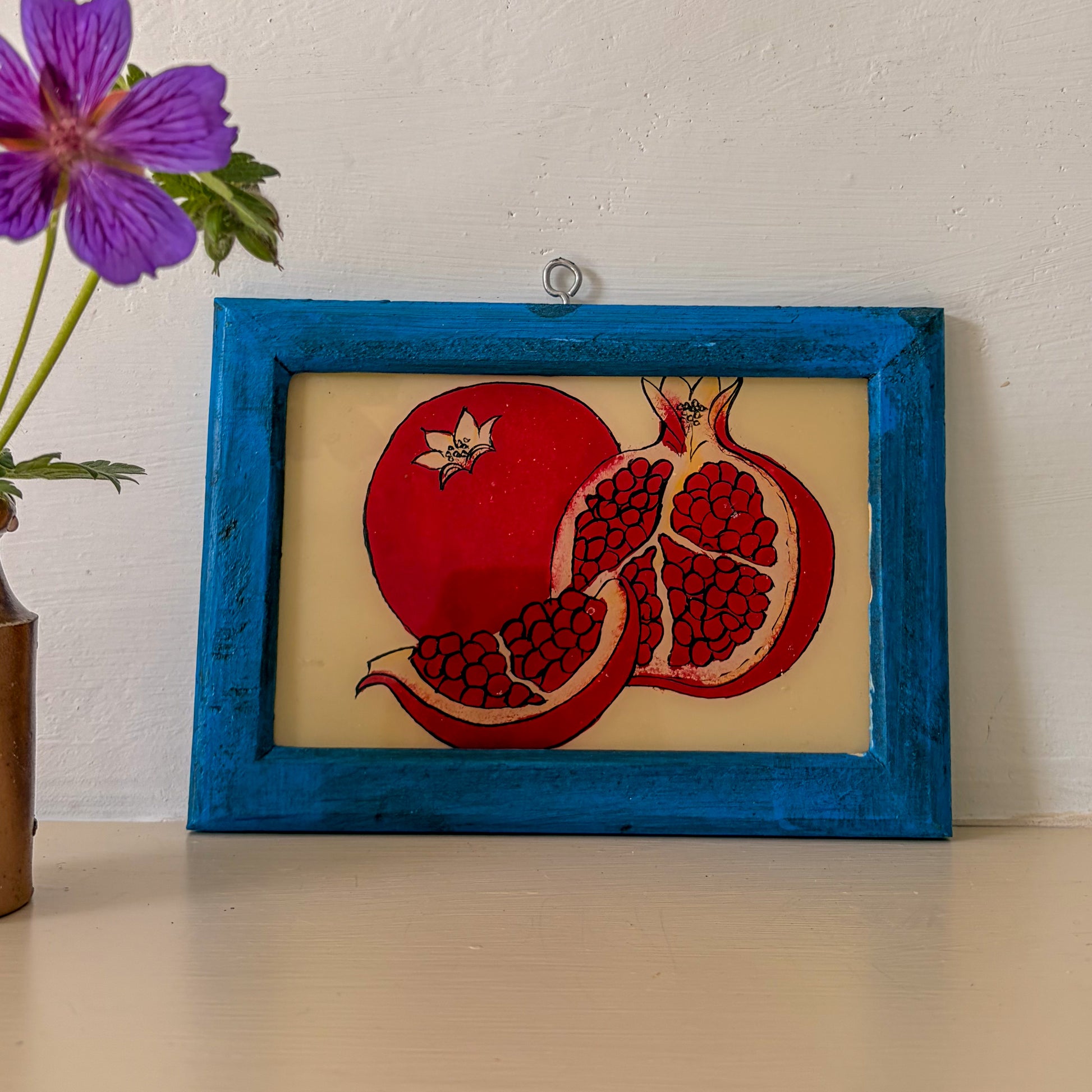 Framed artwork of a pomegranate with a blue frame on a white wall.