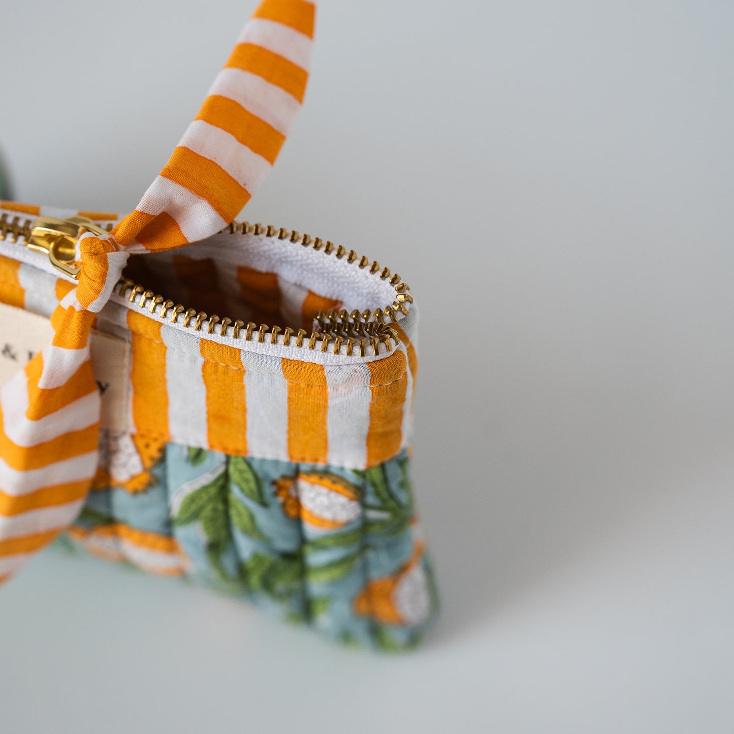 Close-up of a fabric zip pouch with orange and white striped fabric, a matching striped handle, and a green tropical floral patterned base, shown on a light gray background.
