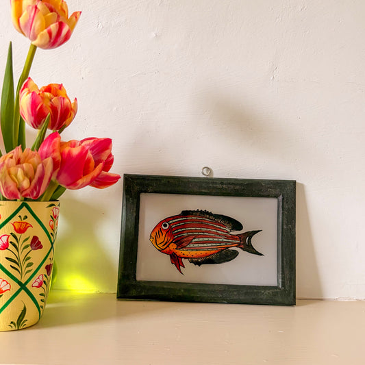 Framed artwork of a fish next to a vase with tulips on a white surface.