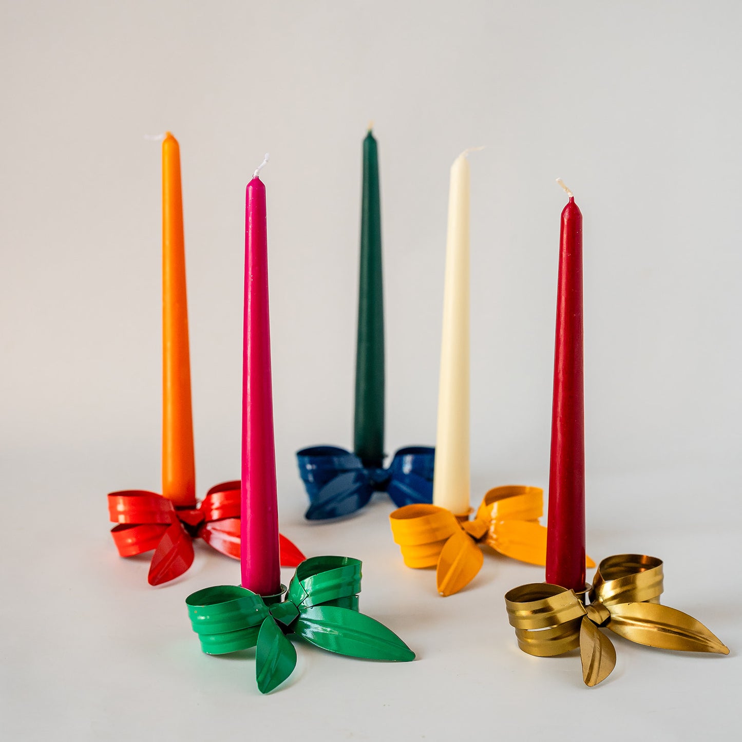 Six taper candles in assorted colors arranged with metallic ribbon bows on a light gray background. The colors include orange, bright pink, dark green, off-white, red, and dark blue, with the bows matching the candle colors.