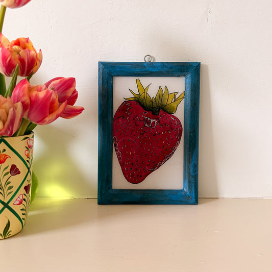 Framed artwork of a strawberry next to a vase of pink tulips on a white surface.