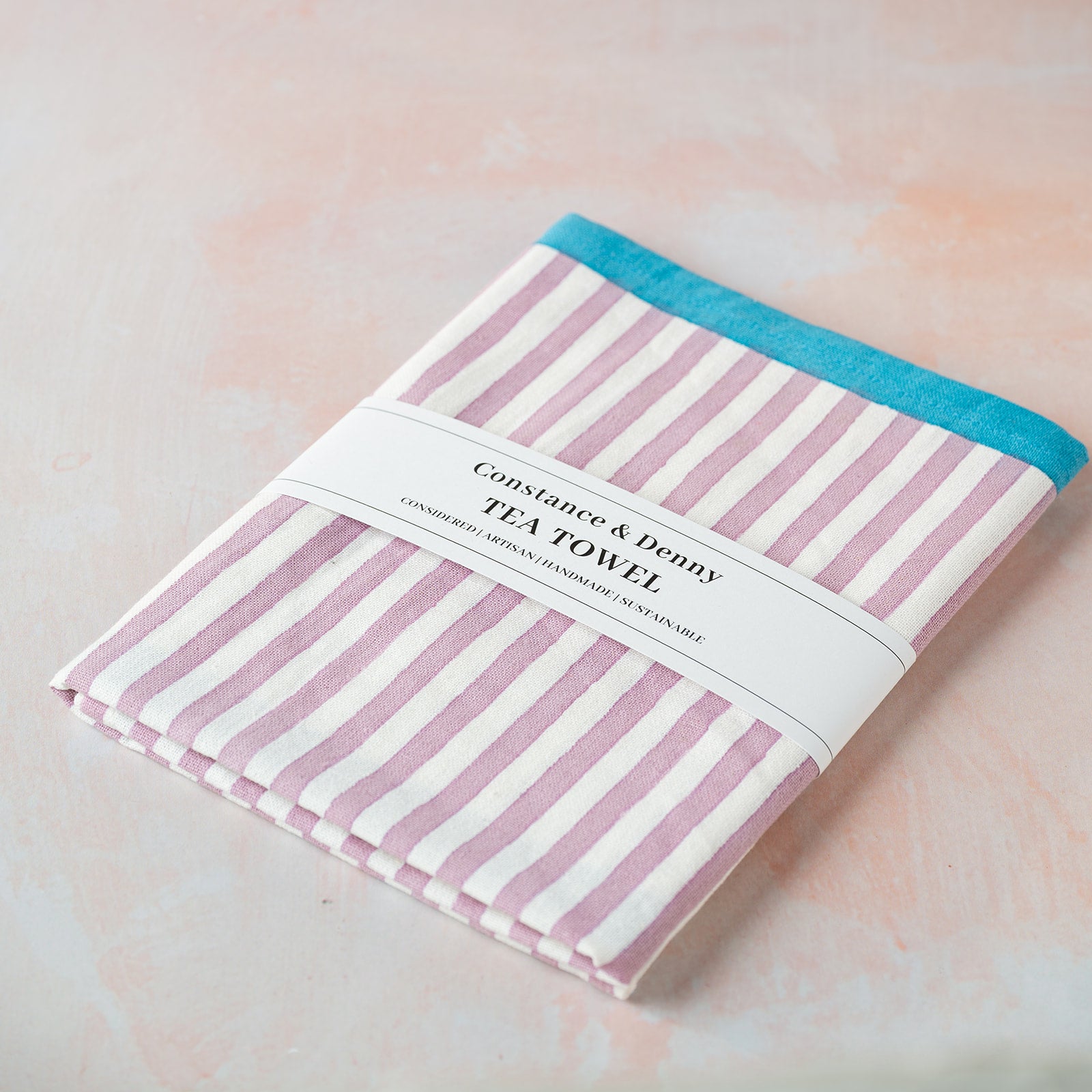 Striped tea towel with pink and white stripes and a blue border on a light pink background.