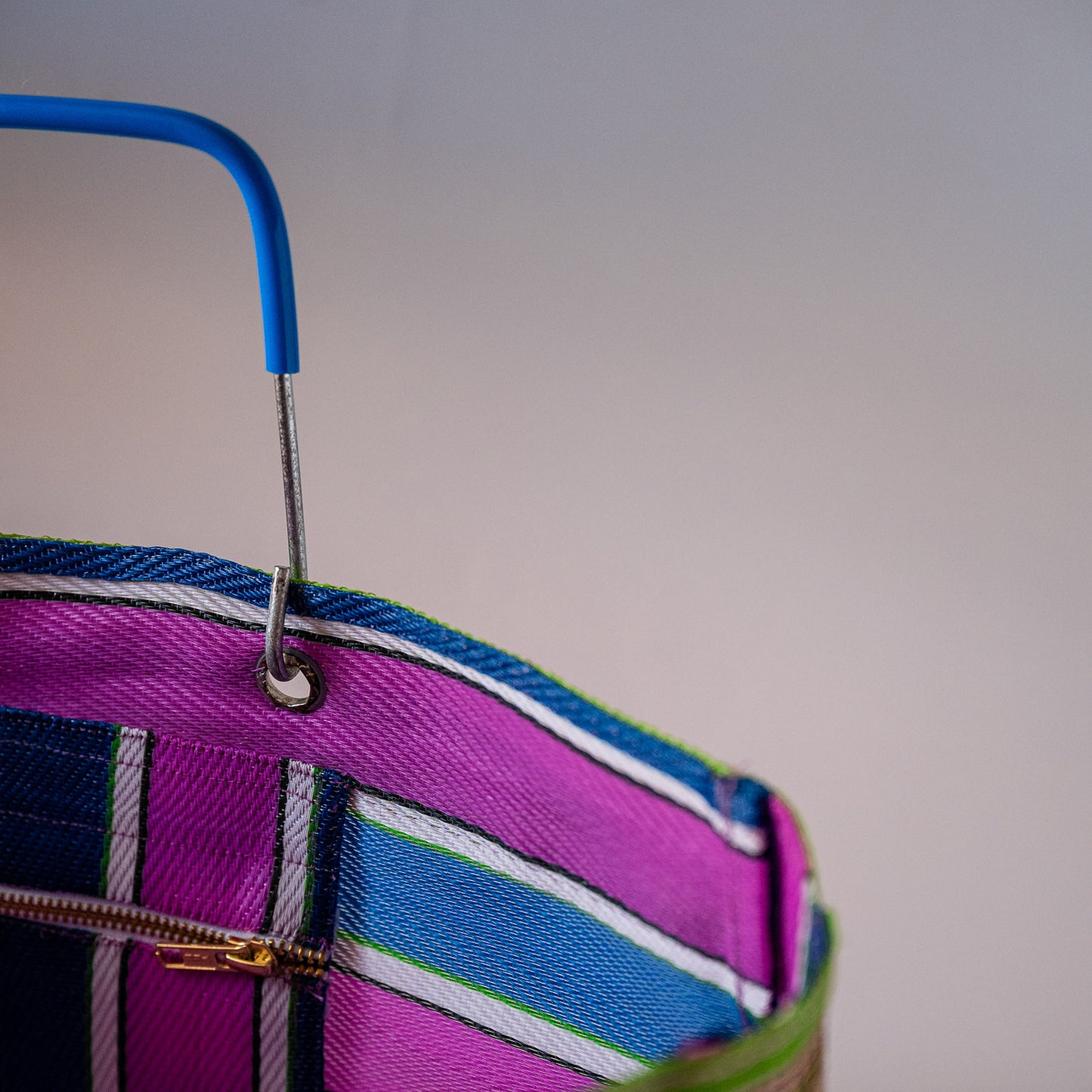Close-up of a colorful striped fabric bag with a bright blue handle and a gold zipper, set against a plain light gray background.