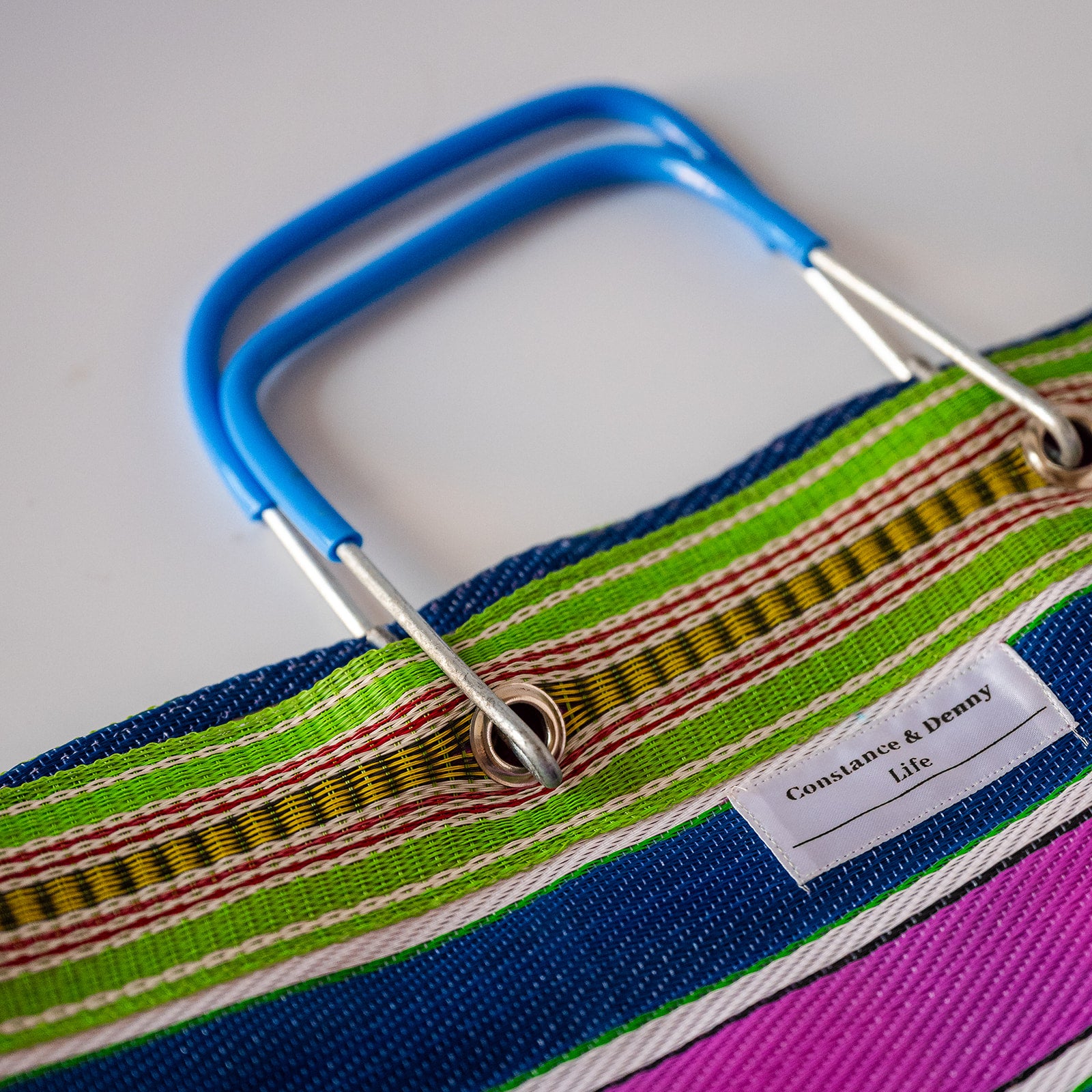 Close-up of a colorful woven tote bag with a blue handle, featuring green, blue, pink, yellow, and red striped fabric with a white brand label reading 'Constantine & Denny Life'.