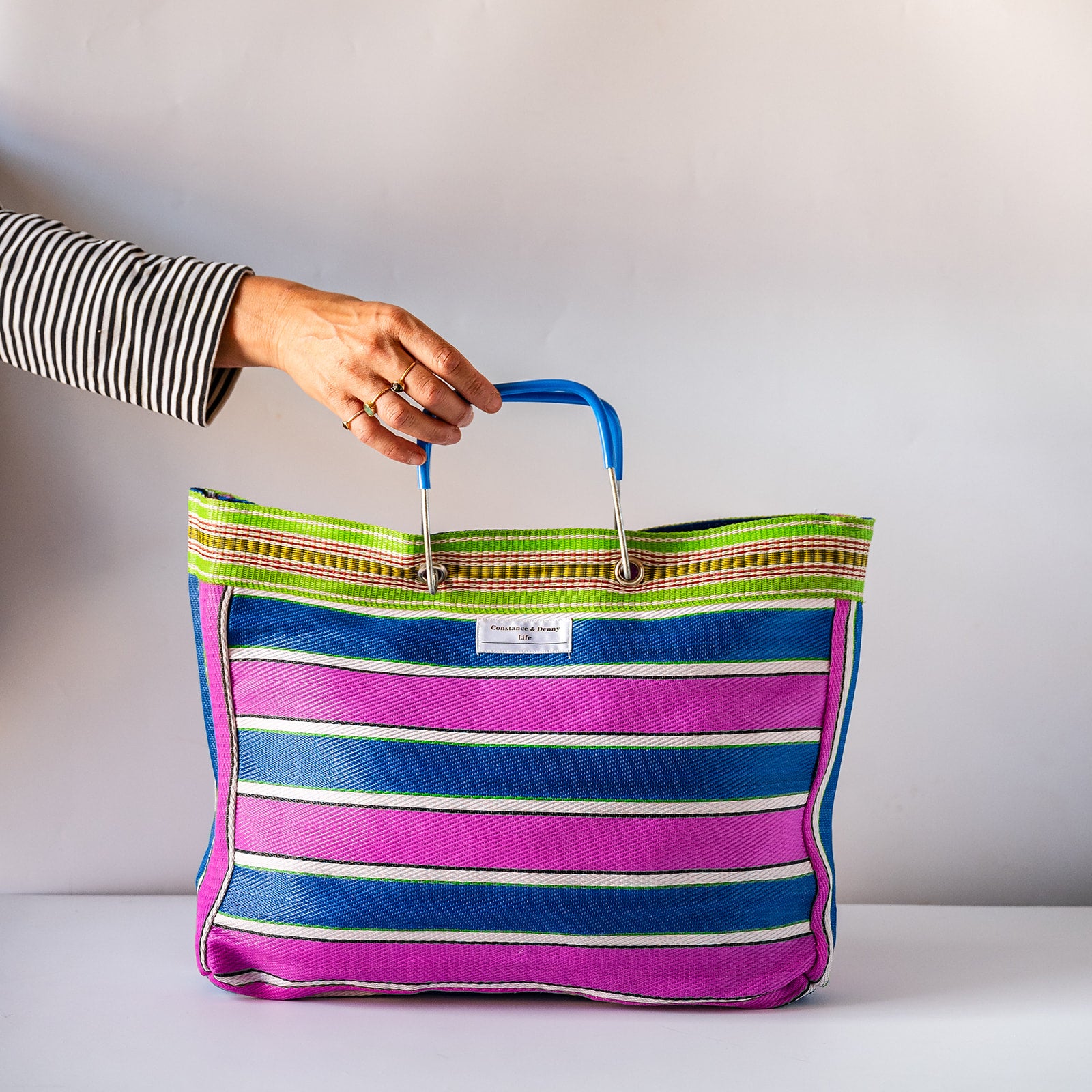A striped woven tote bag with pink, blue, and white horizontal stripes, held by a person's hand. The bag features a green and red patterned top trim and a white brand label that reads 'Tote Bag'.
