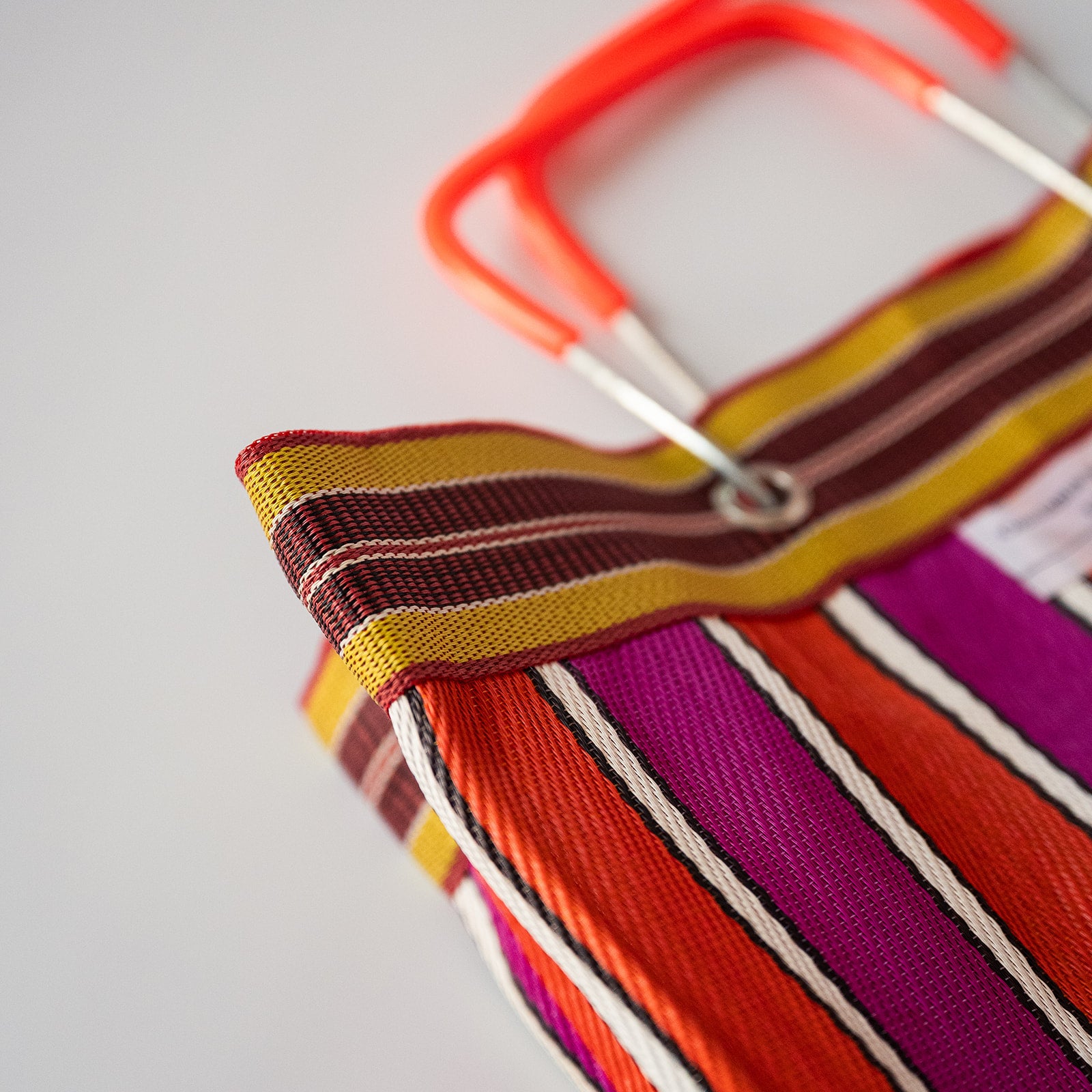 Close-up of a colorful striped woven bag with orange, red, purple, yellow, and brown stripes, featuring a bright orange handle and silver metal hardware on a light gray background.
