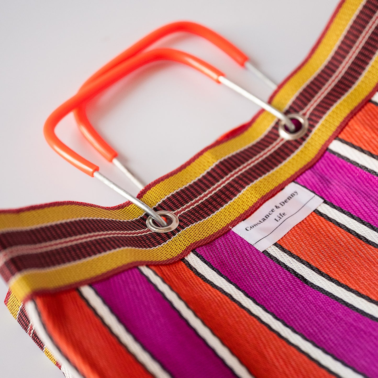 Close-up of a striped tote bag with bright orange handles, featuring bold stripes in orange, magenta, yellow, brown, and white. A small fabric label reads 'Conscious Living'.